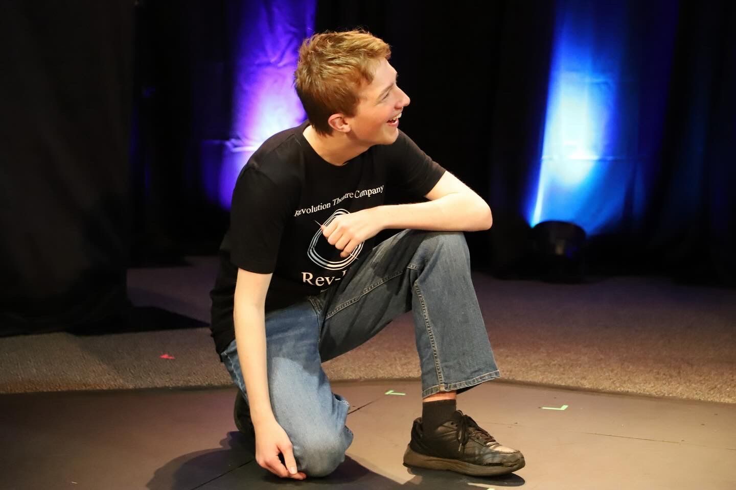 A young man is kneeling down on a stage in front of a blue light.