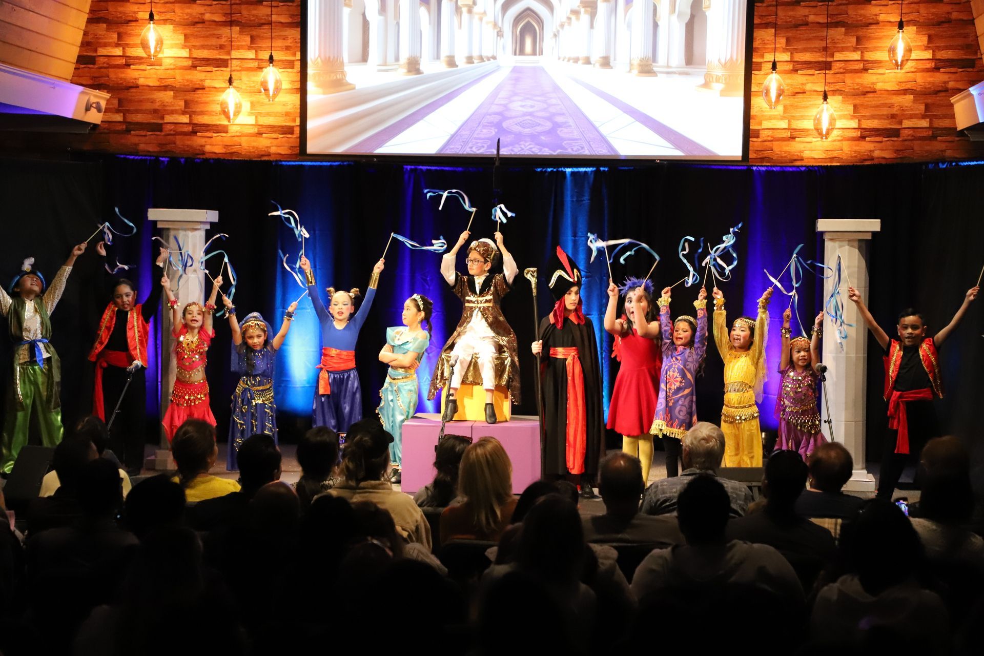 A group of children are dancing on a stage in front of a large screen.