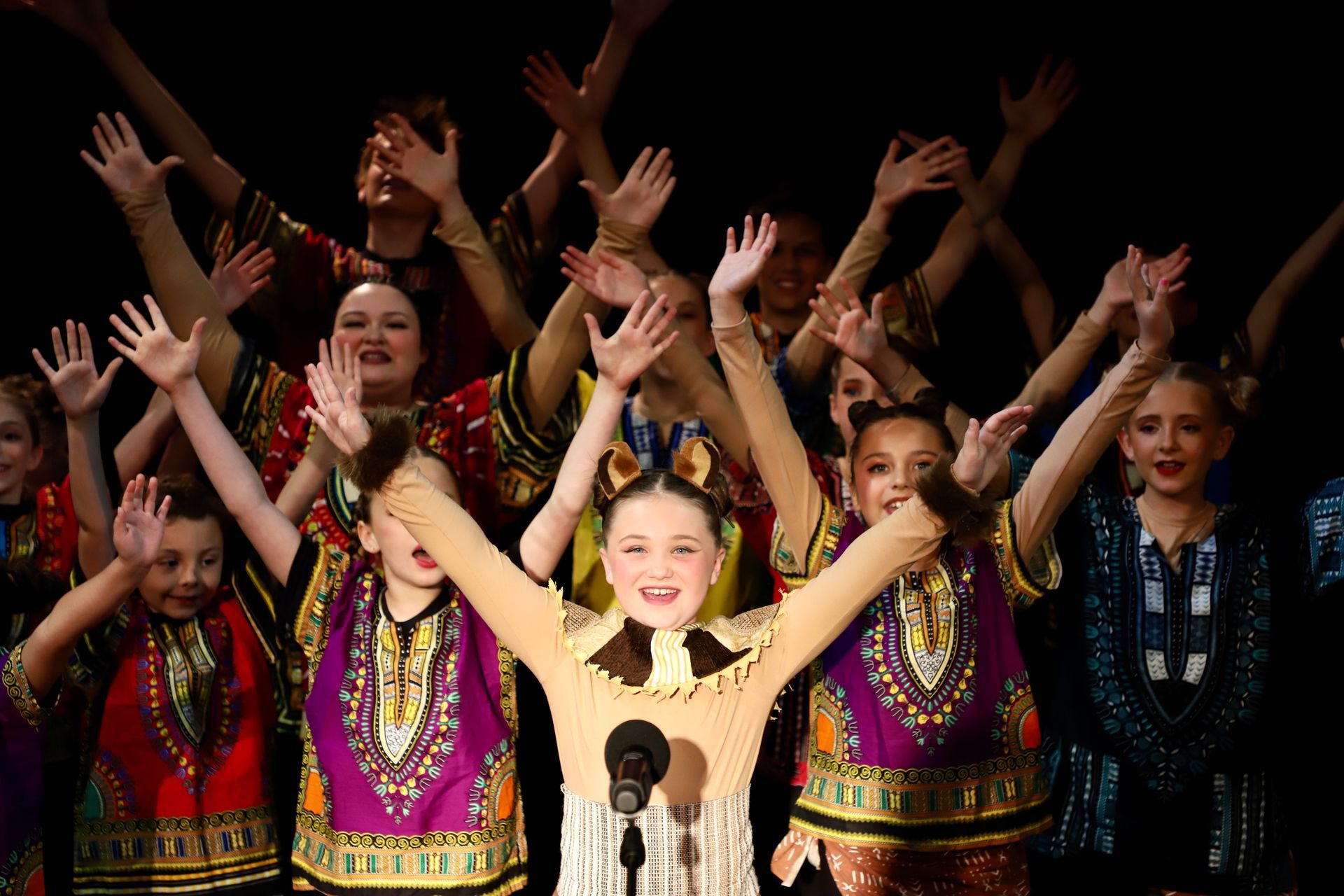 A girl is singing into a microphone in front of a crowd of children.