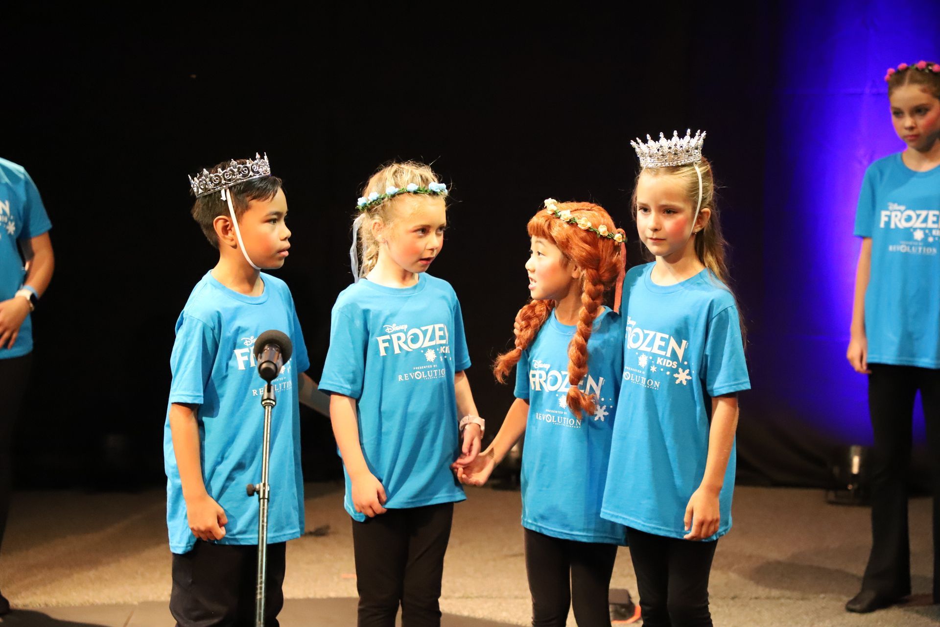 A group of children wearing blue shirts with the word frozen on them are standing next to each other on a stage.