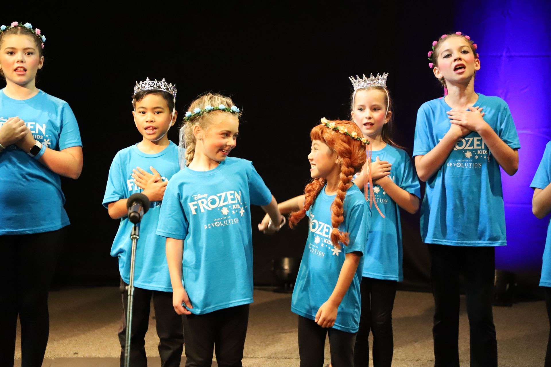 A group of young girls wearing blue frozen shirts are singing into microphones.