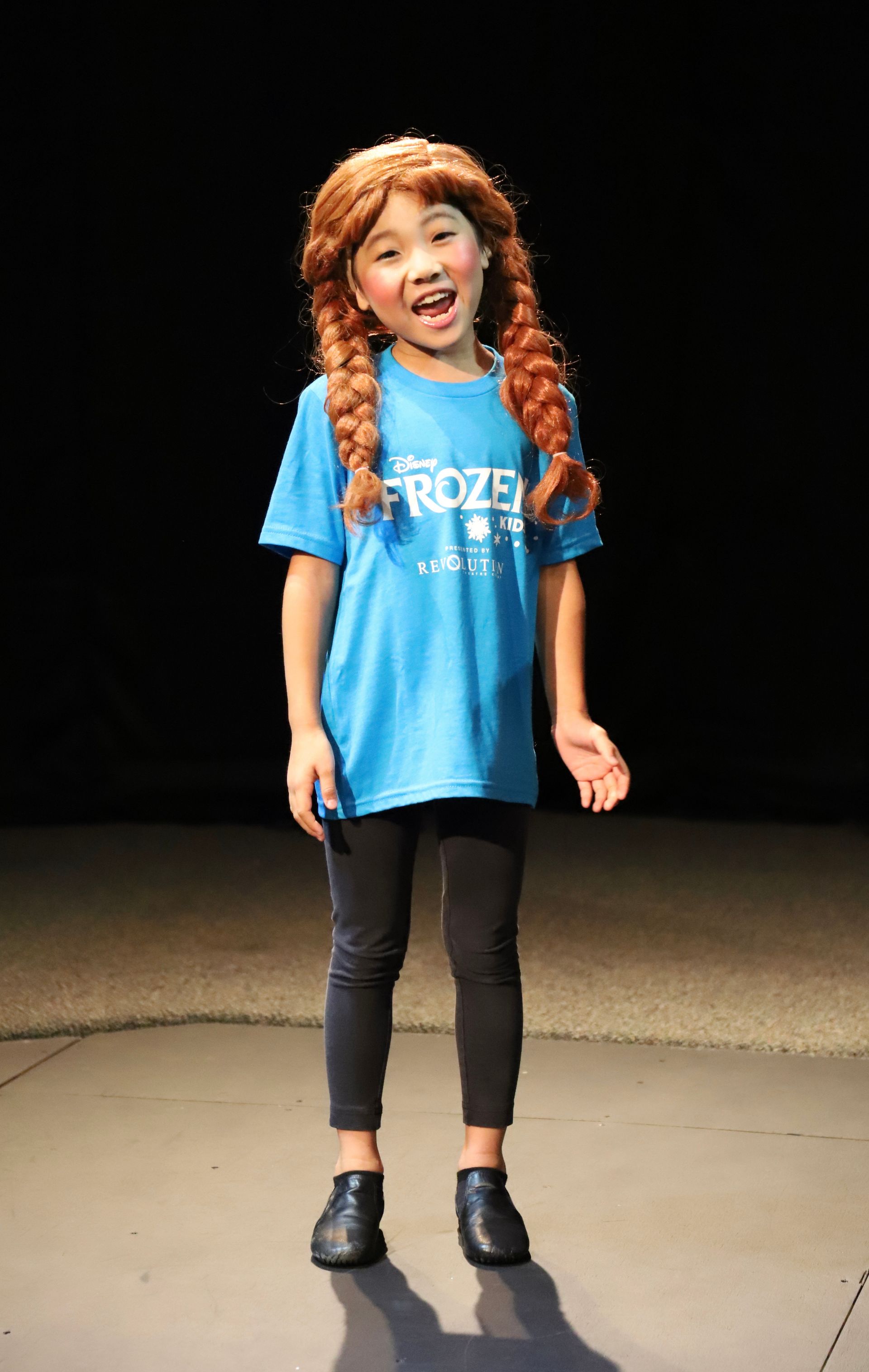 A young girl wearing a wig and a blue shirt is standing on a stage.