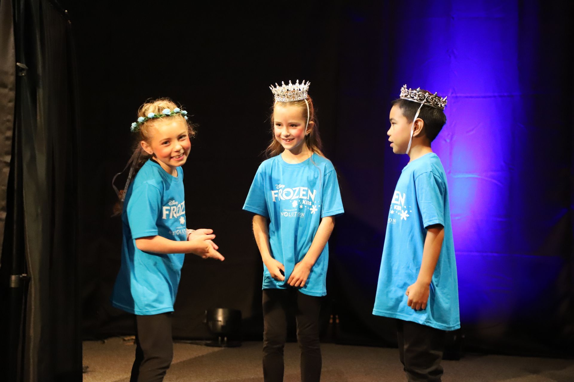 Three children wearing blue shirts and crowns are standing on a stage.