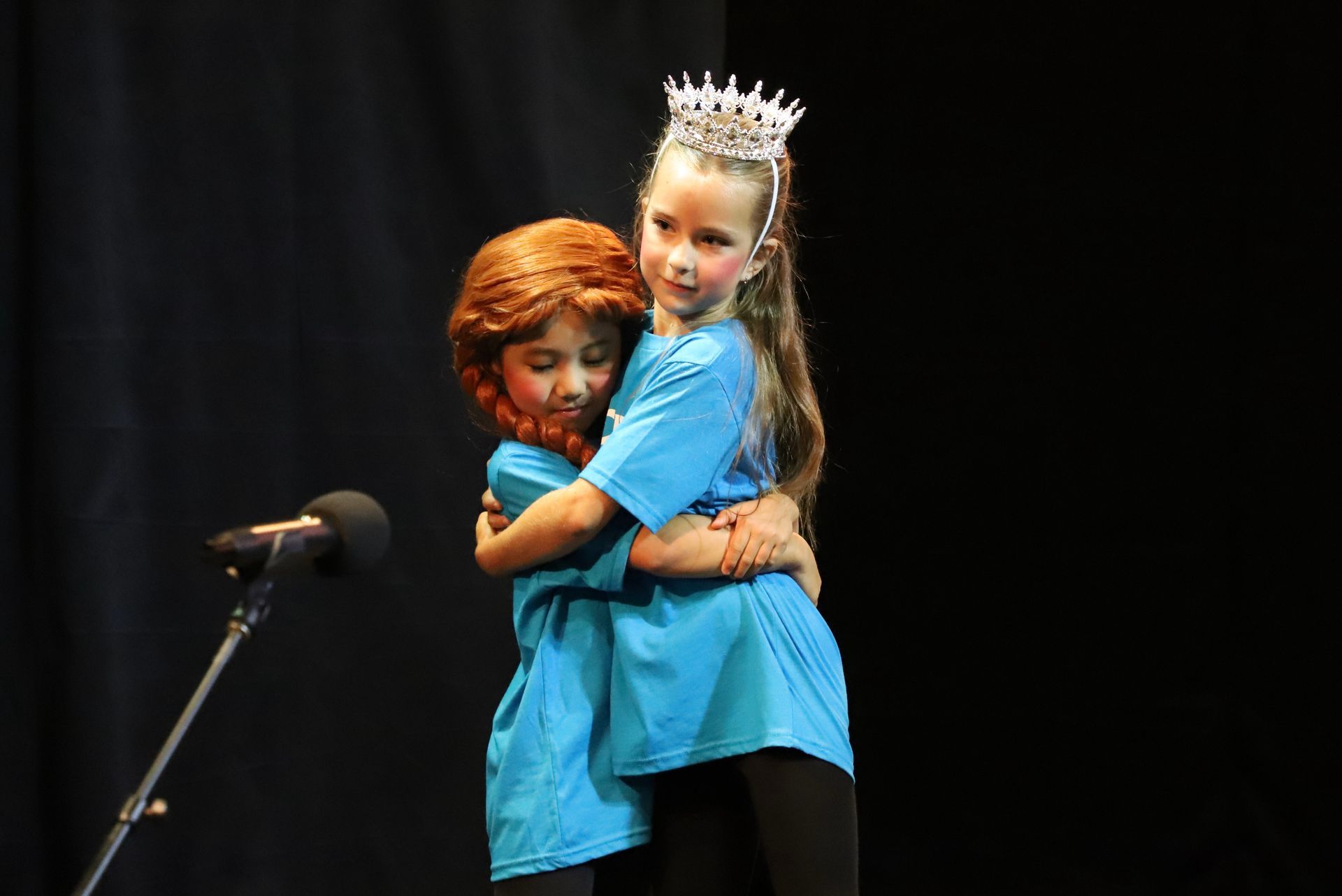 Two little girls are hugging each other on a stage in front of a microphone.