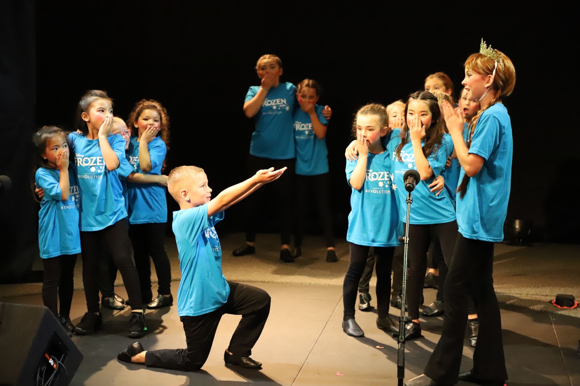 A group of children in blue shirts are singing into microphones on a stage.