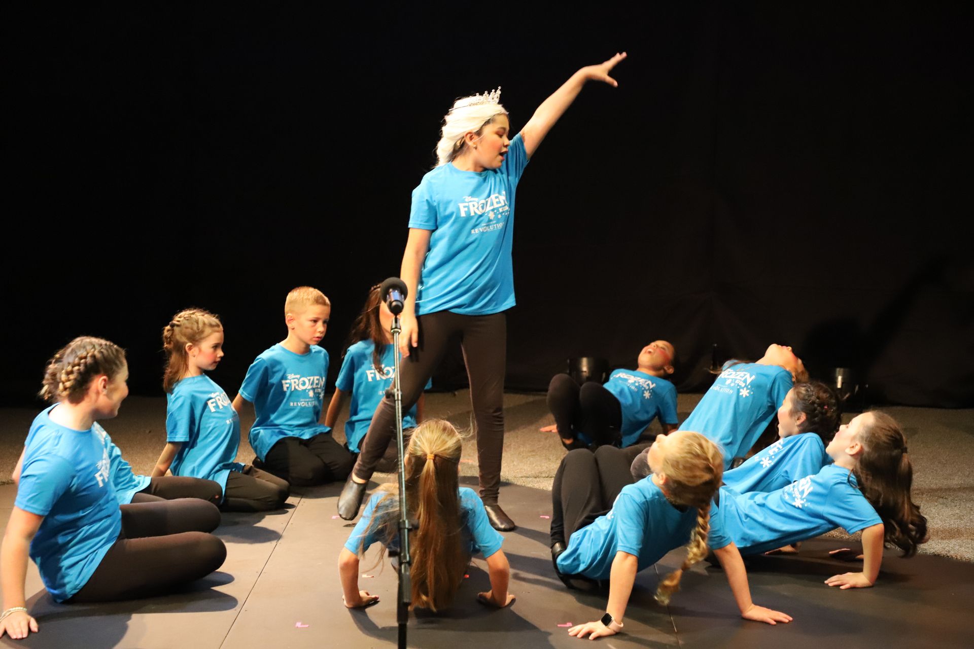 A group of children in blue shirts are sitting on the floor on a stage.