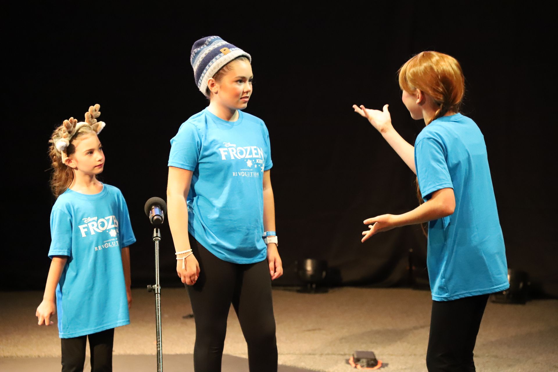 Three children wearing blue shirts that say frozen are standing on a stage.