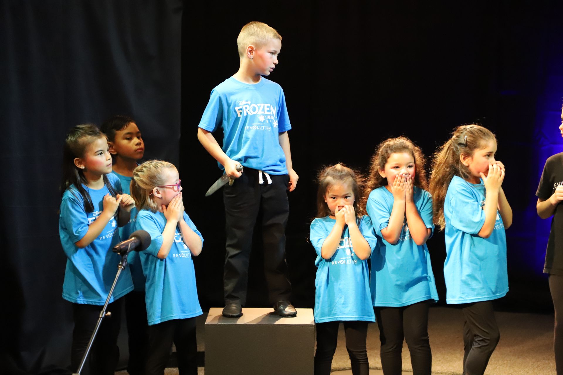A group of children in blue shirts are singing into microphones on a stage.
