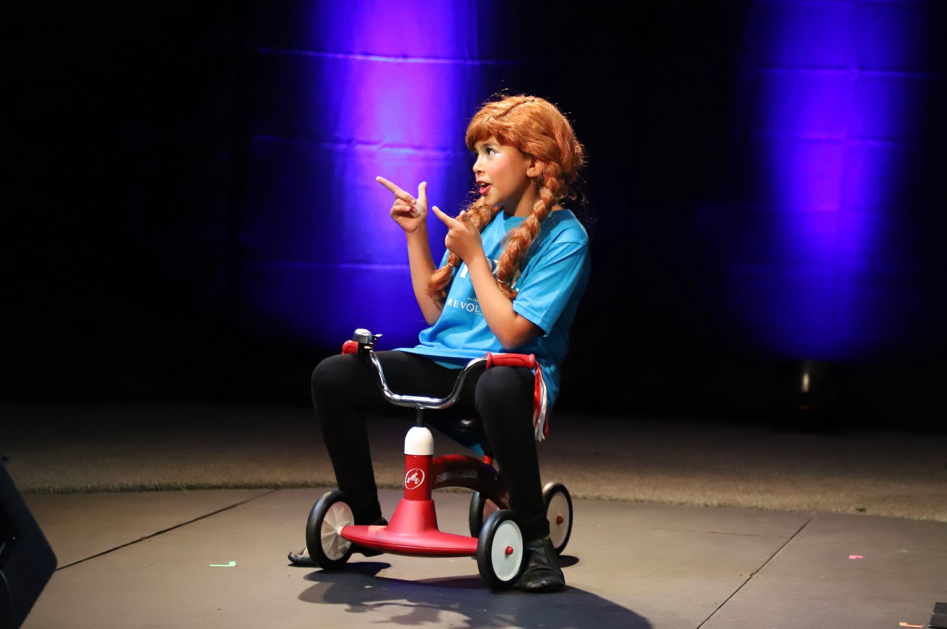 A young girl is sitting on a red tricycle on a stage.