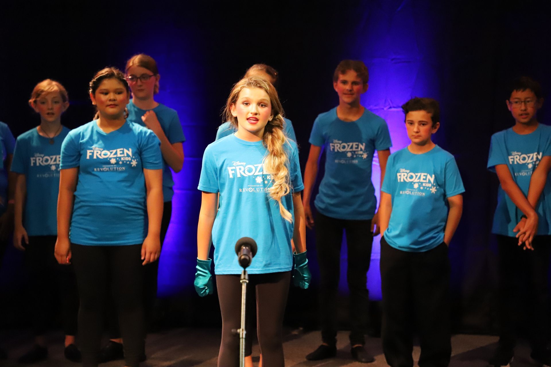 A girl is singing into a microphone in front of a group of children wearing blue shirts.