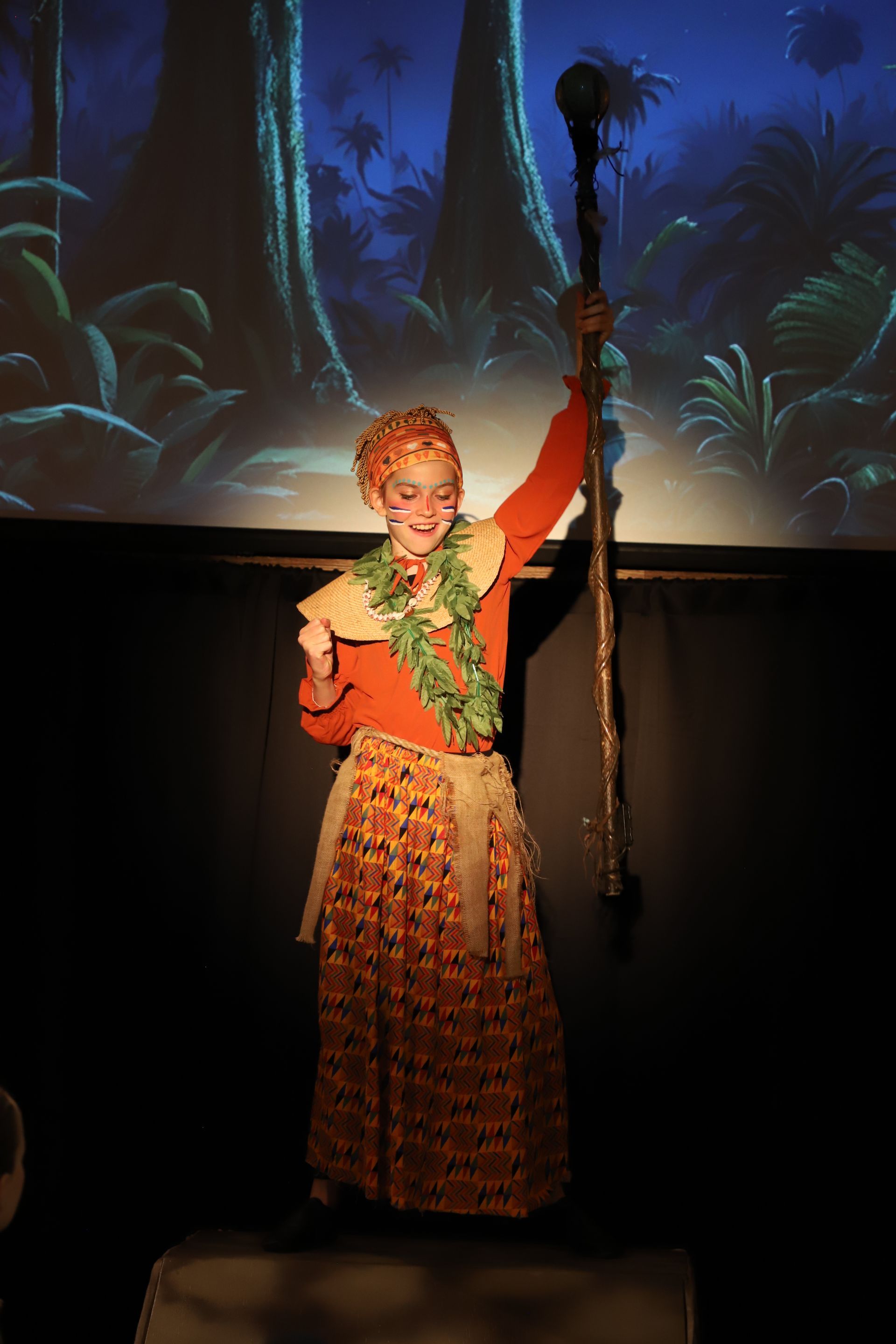 A woman in a costume is holding a cane in front of a screen.