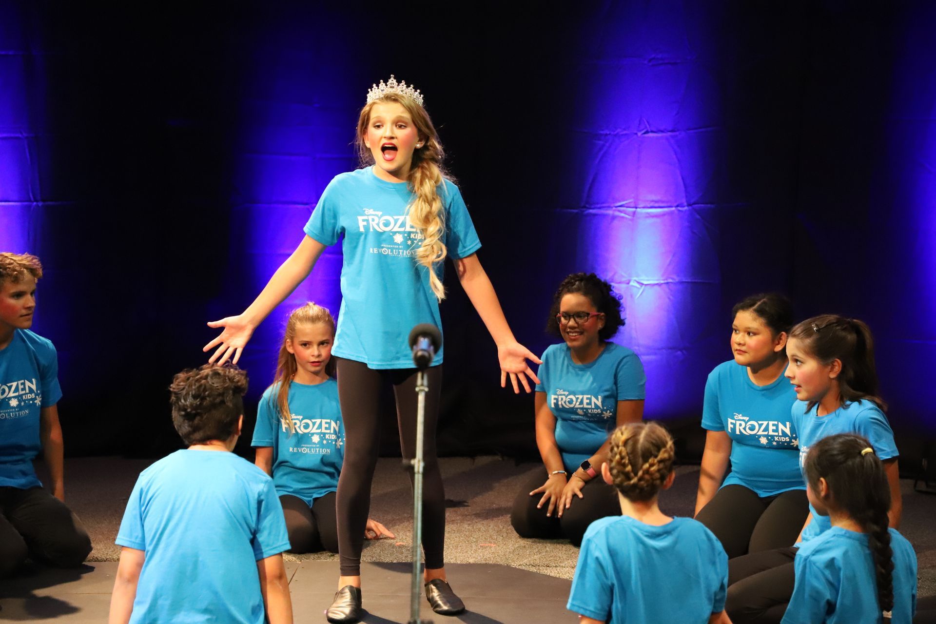 A girl is singing into a microphone in front of a group of children.