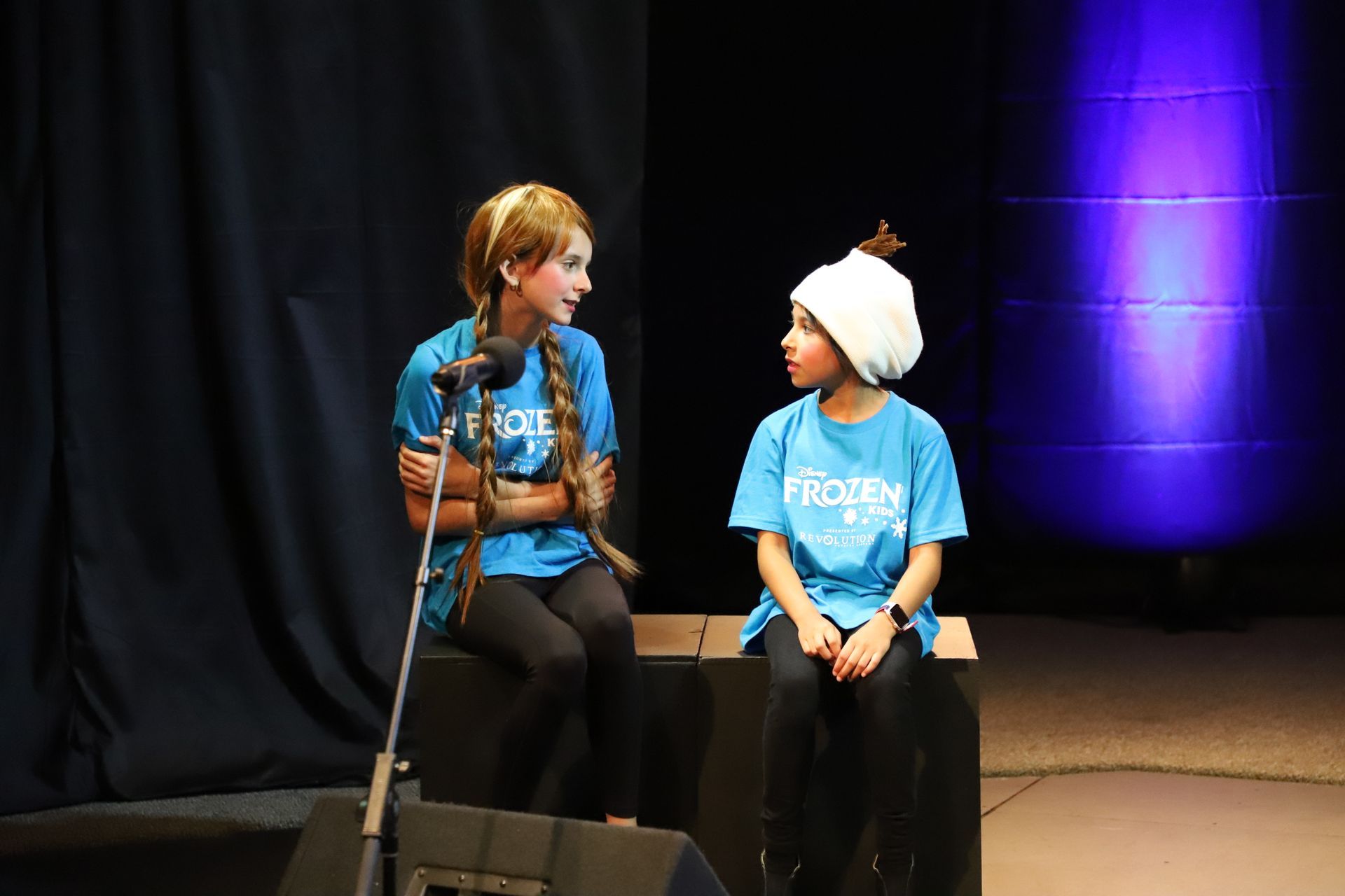 Two children wearing frozen shirts are sitting next to each other on a stage