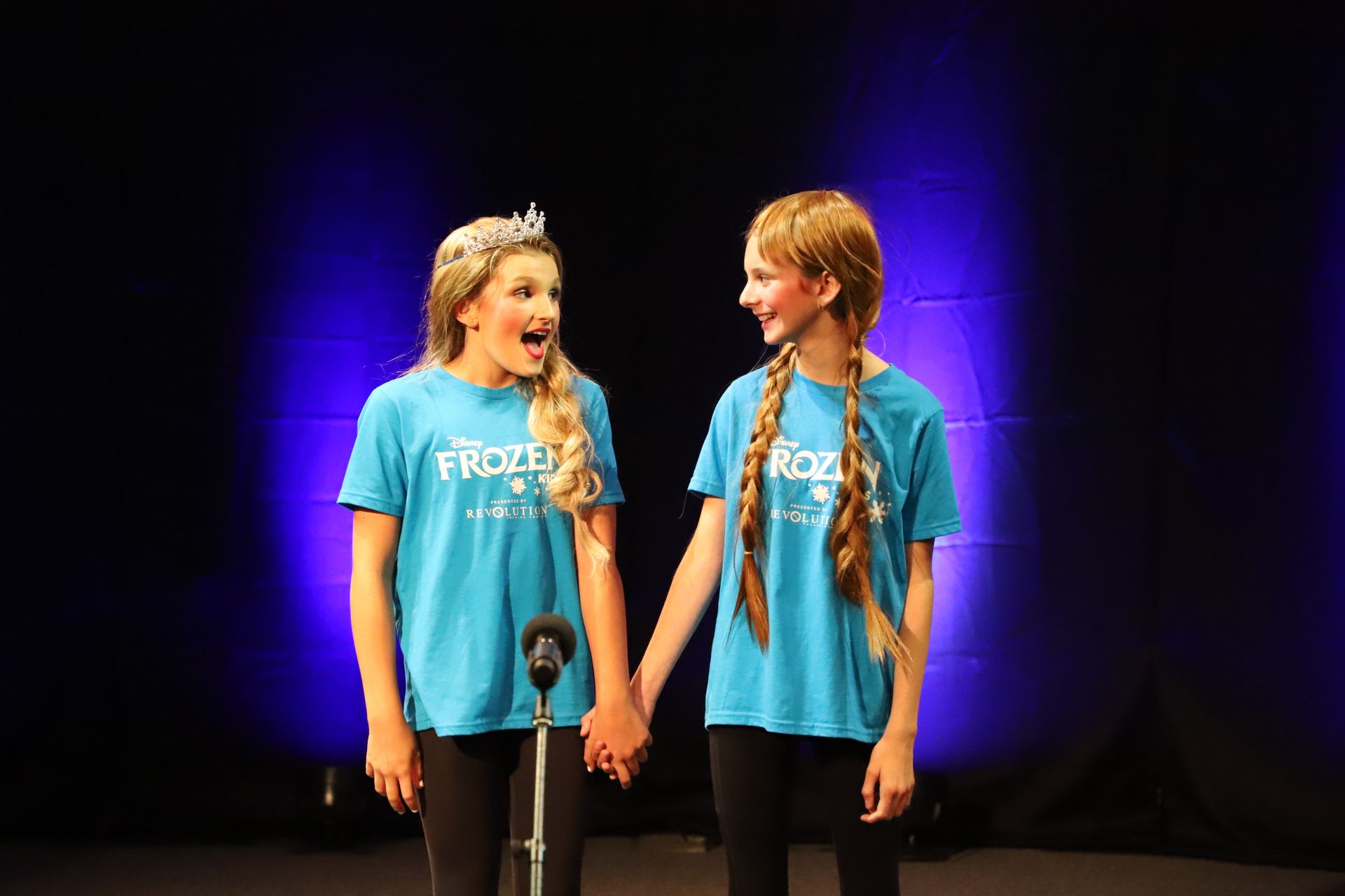 Two young girls are holding hands and singing into microphones on a stage.