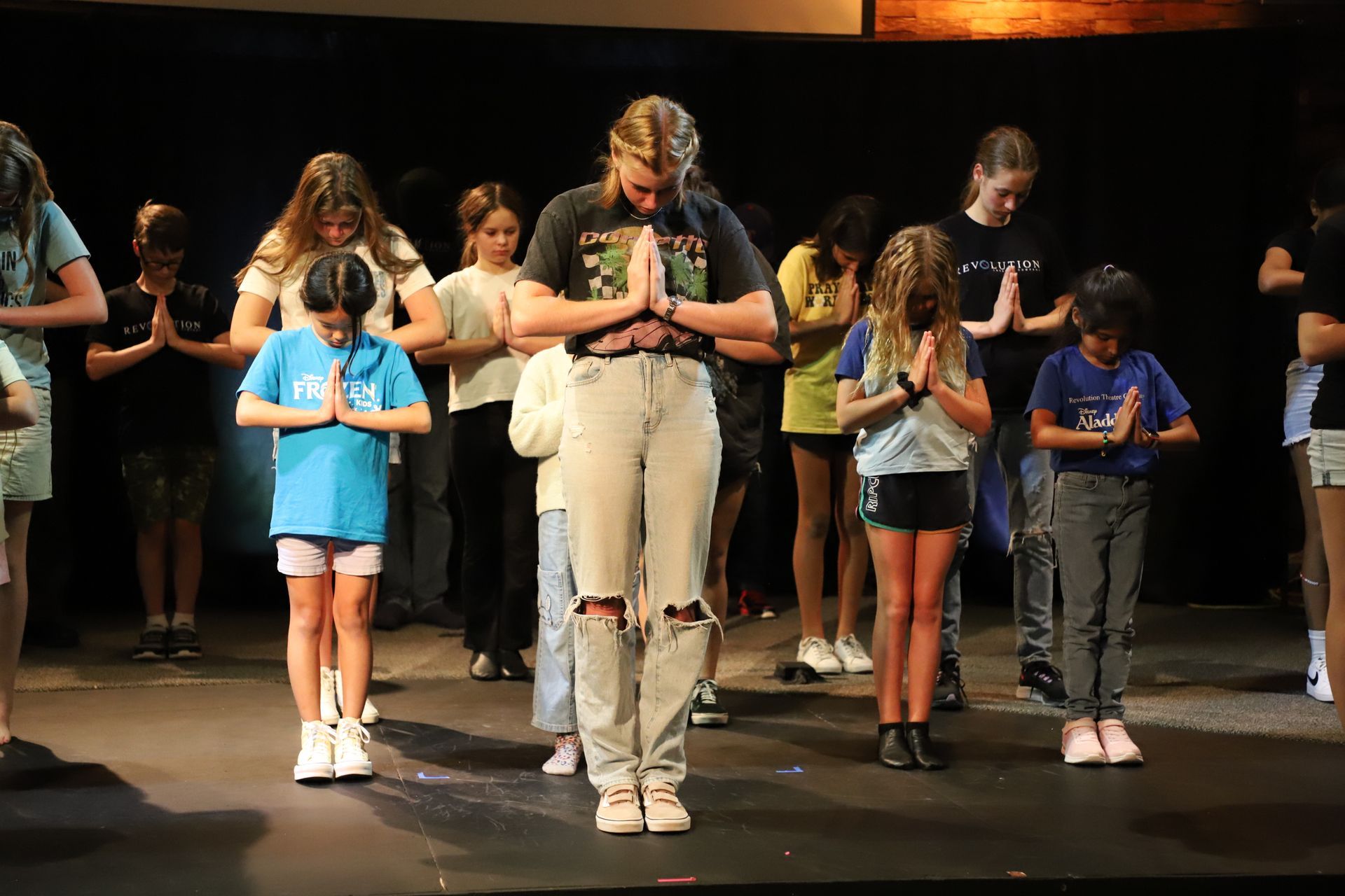 A group of children are standing on a stage with their hands folded in prayer.