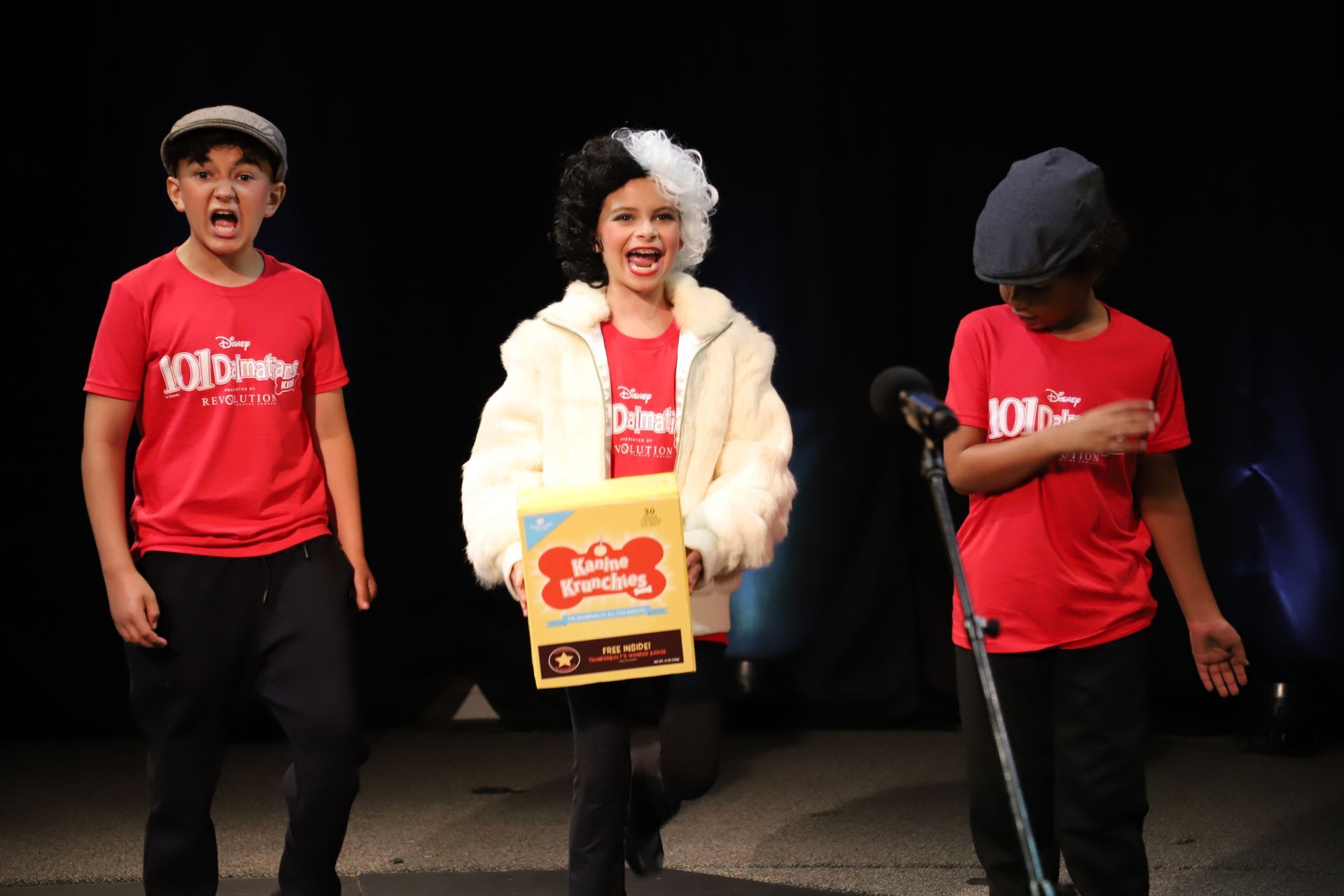 Three children are standing on a stage holding a box of dog treats.