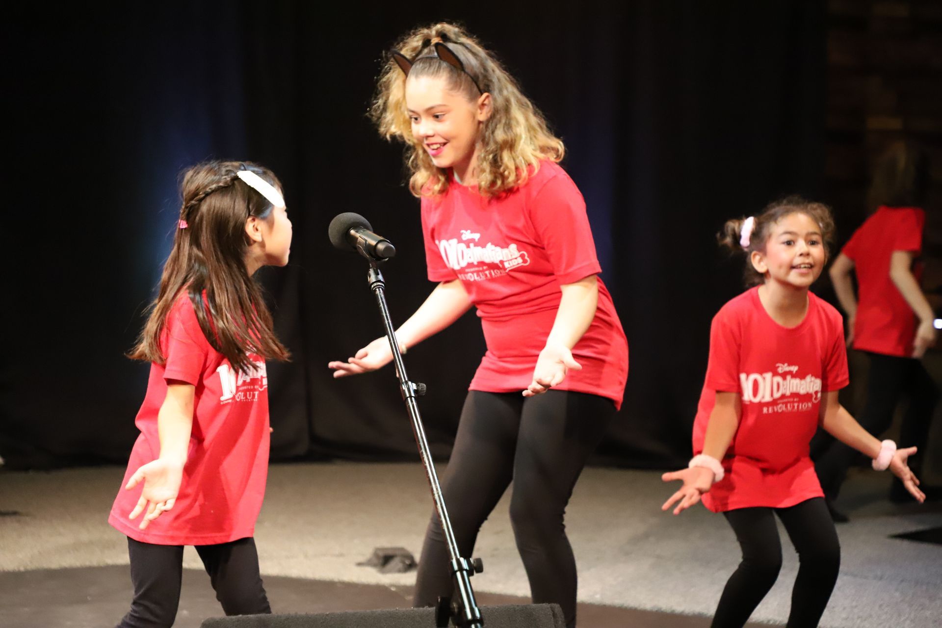 A girl in a red shirt is singing into a microphone while two other girls are dancing.