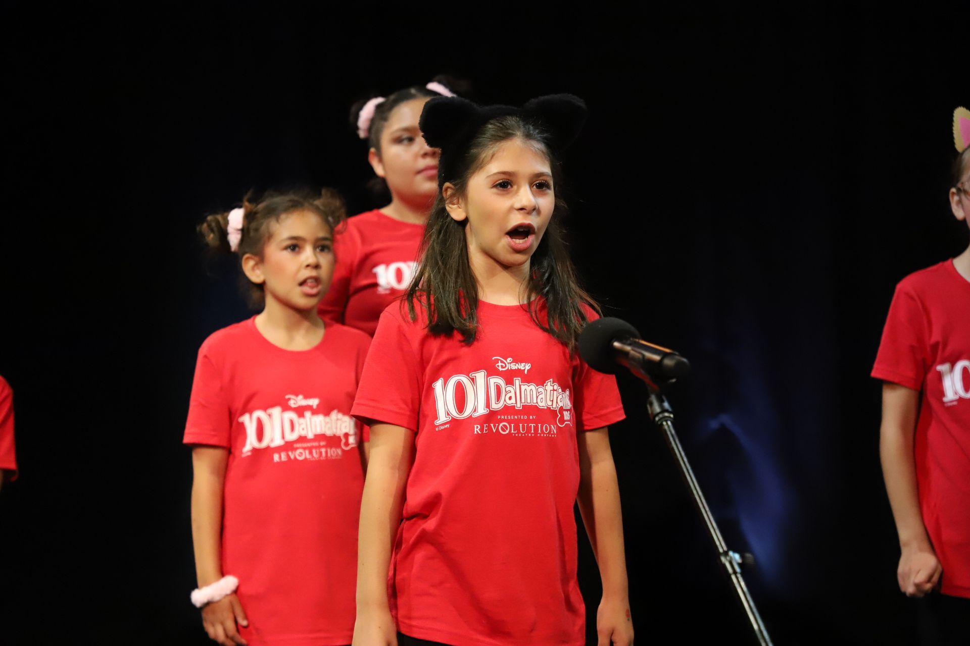 A group of young girls in red shirts are singing into a microphone.