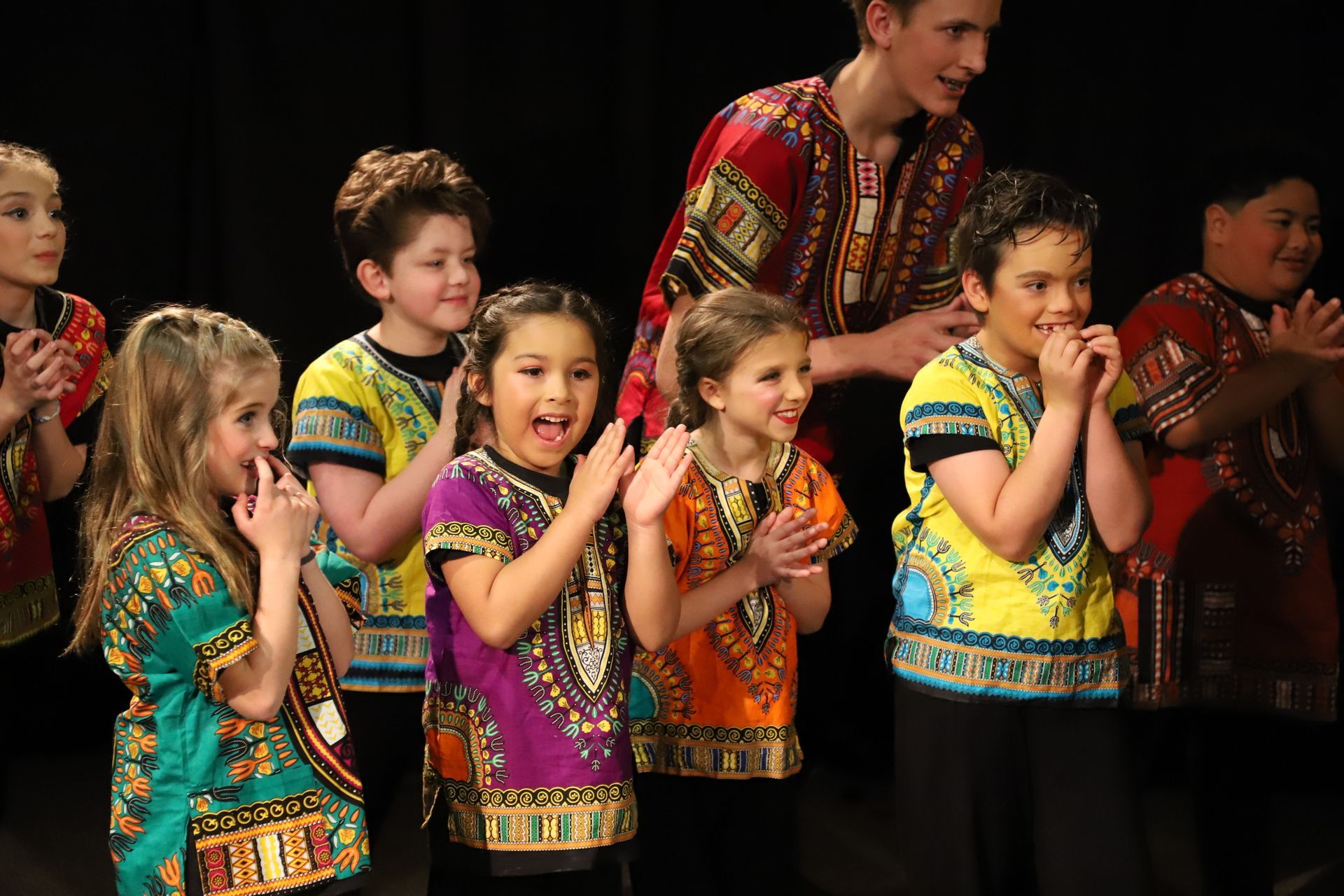 A group of children in colorful shirts are clapping their hands on a stage.
