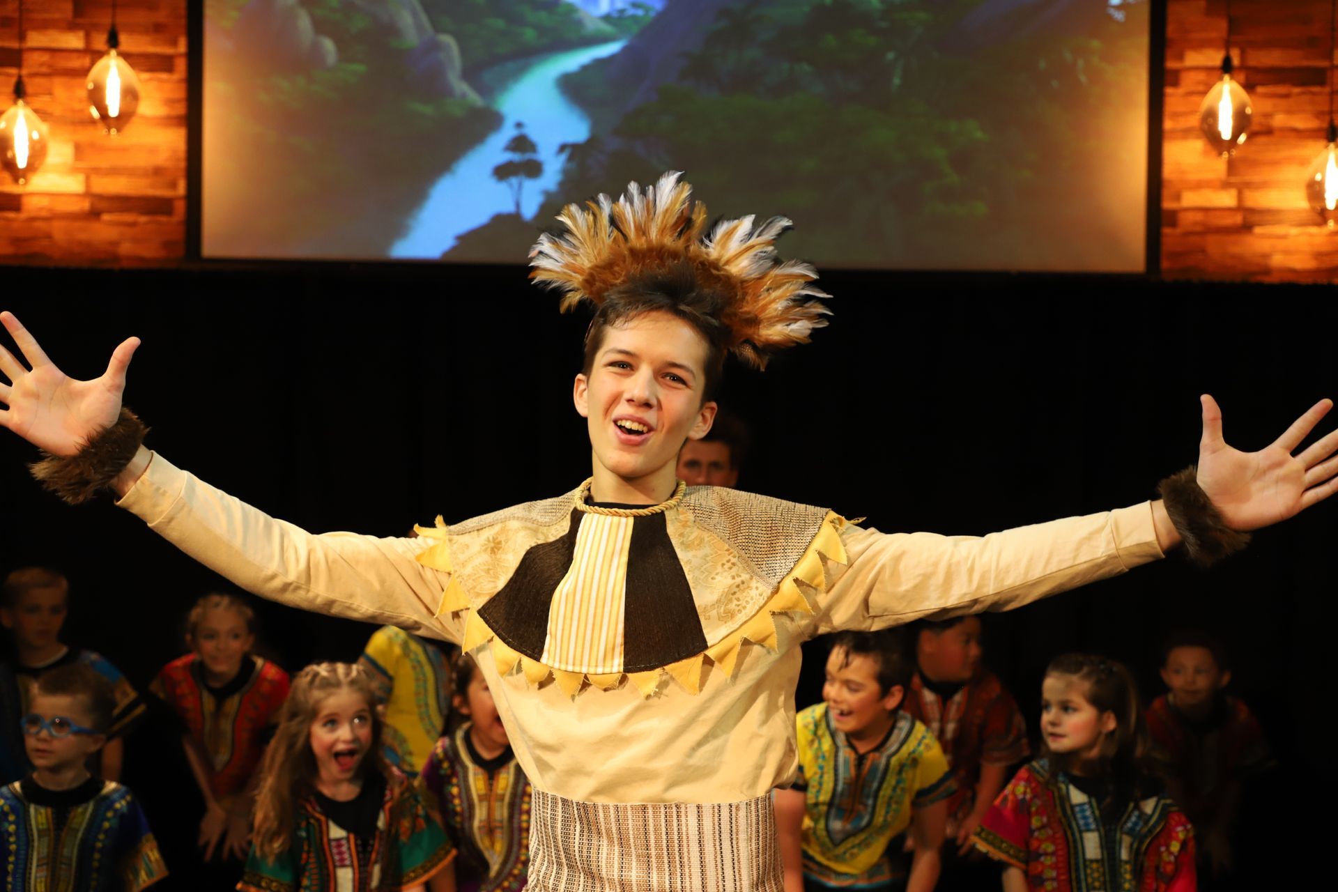 A boy in a native american costume is standing on a stage with his arms outstretched.