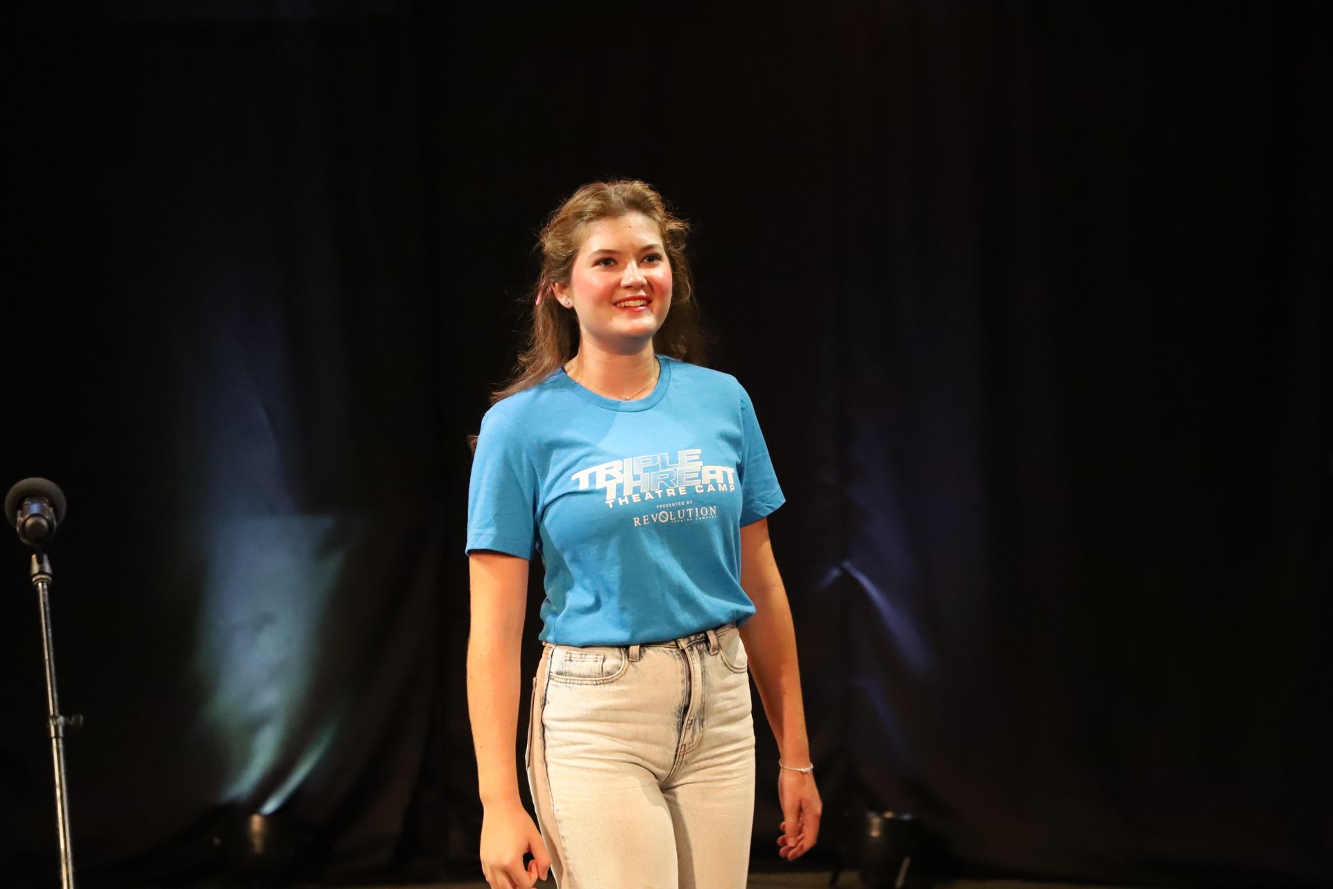 A woman in a blue shirt is standing in front of a microphone on a stage.