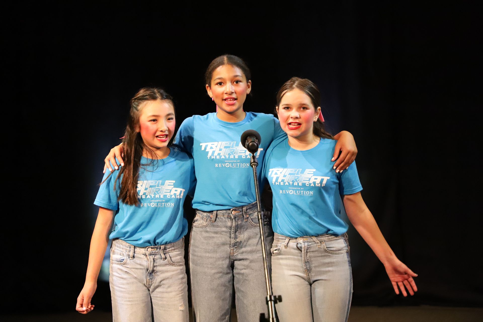 Three young girls are singing into microphones on a stage.