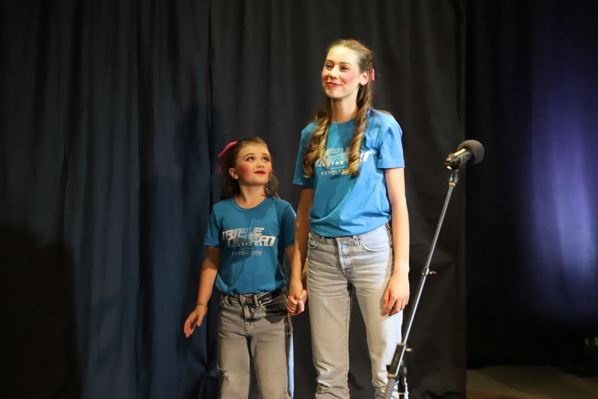 Two young girls are standing next to each other on a stage in front of a microphone.