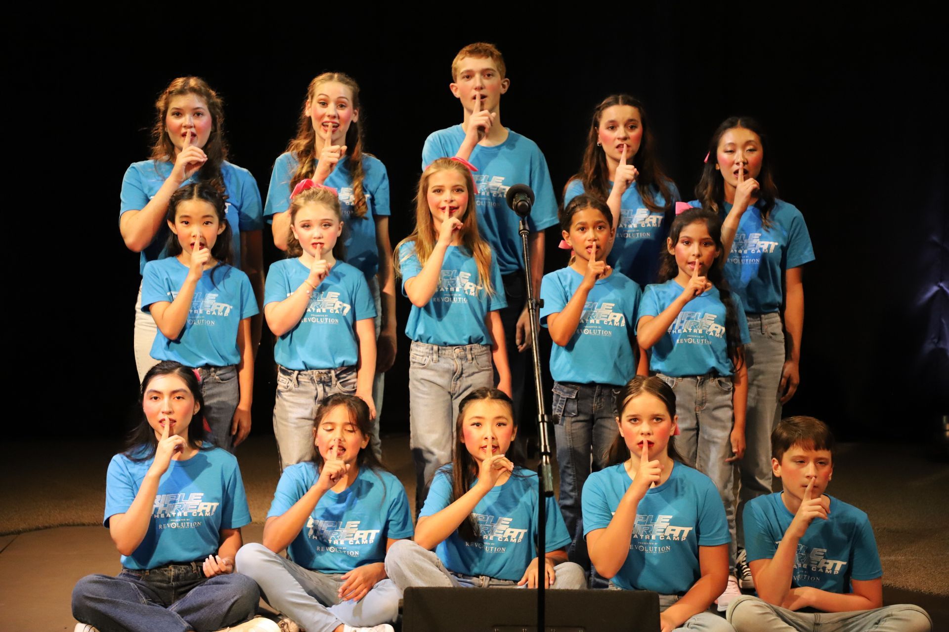 A group of young people are sitting on the floor with their hands to their mouths.