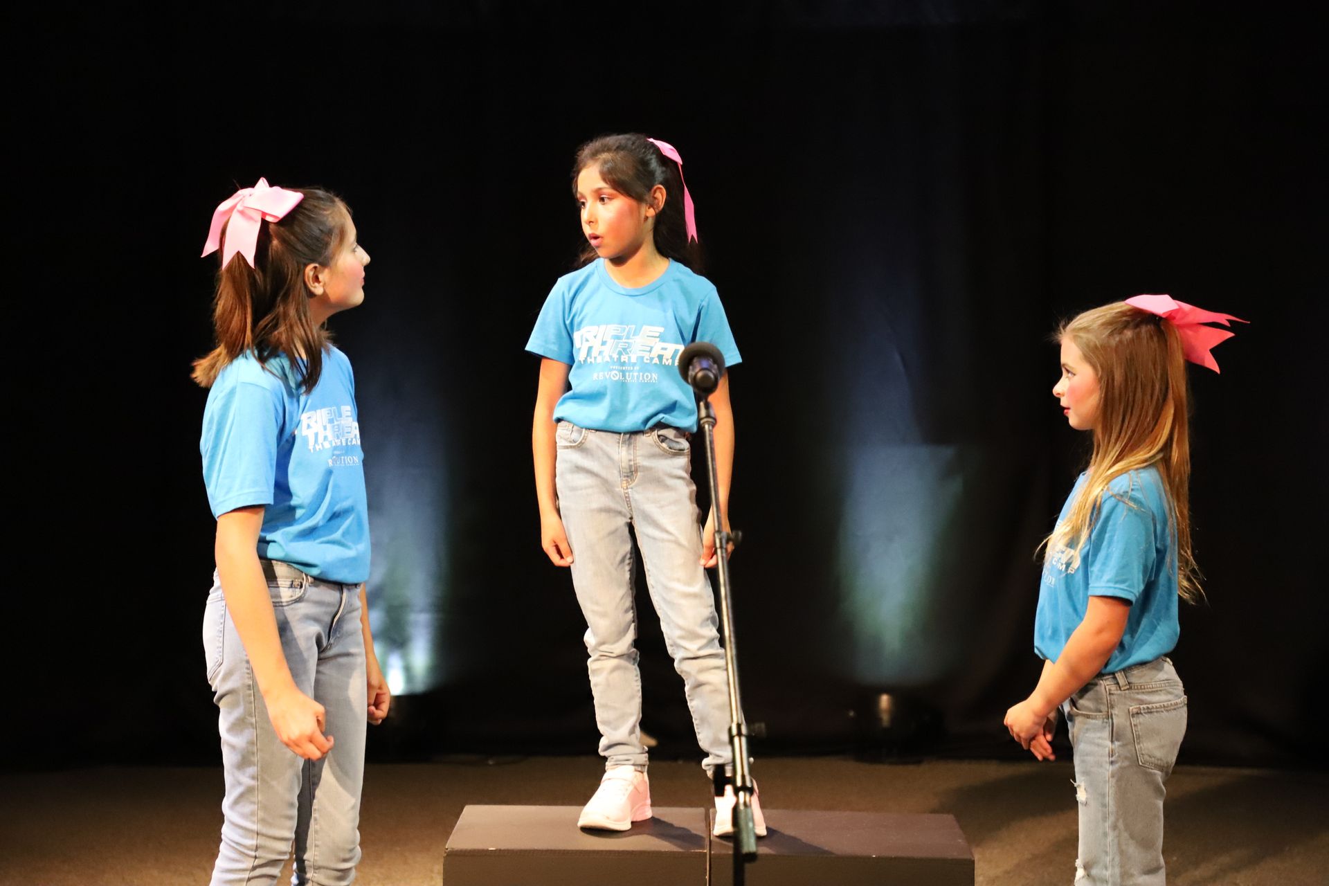 Three young girls are standing in front of a microphone on a stage.