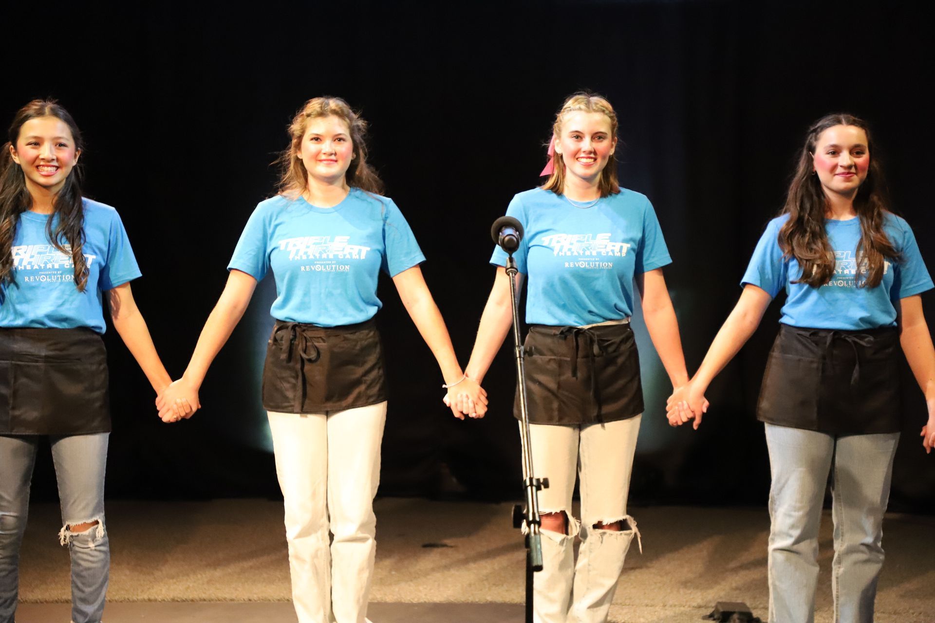 Four young women are holding hands in front of a microphone on a stage.