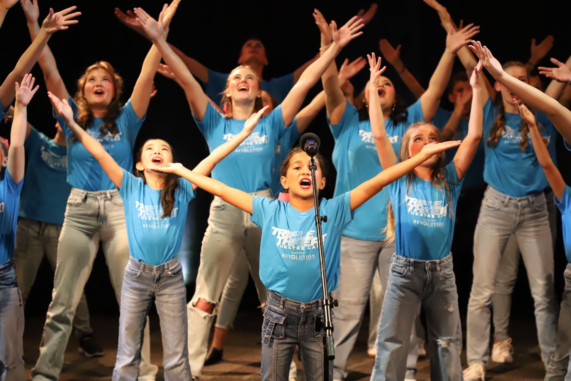 A group of young girls in blue shirts are singing into a microphone.