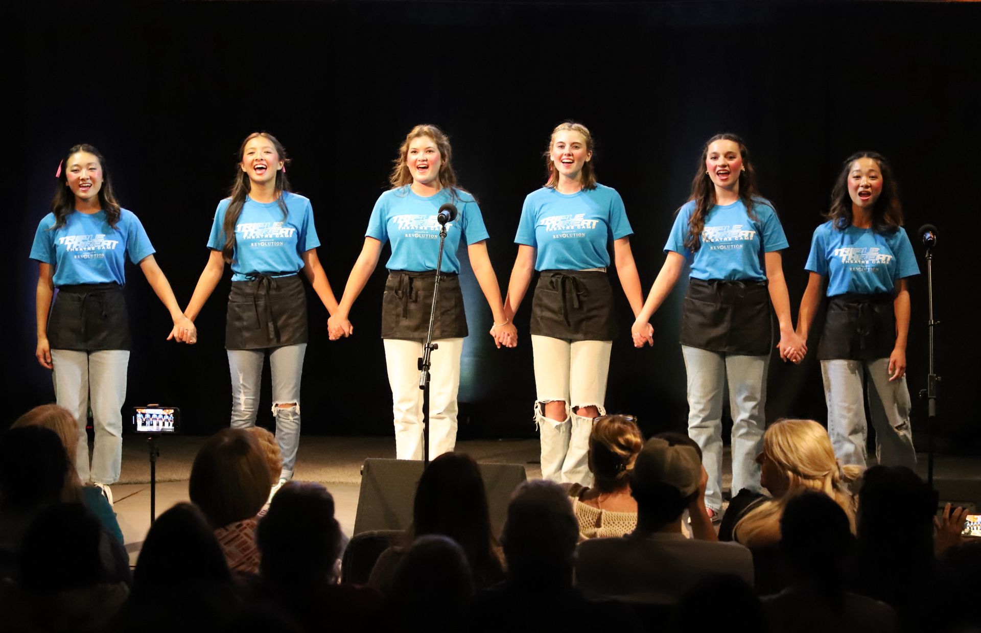 A group of young women are holding hands on a stage in front of a crowd.
