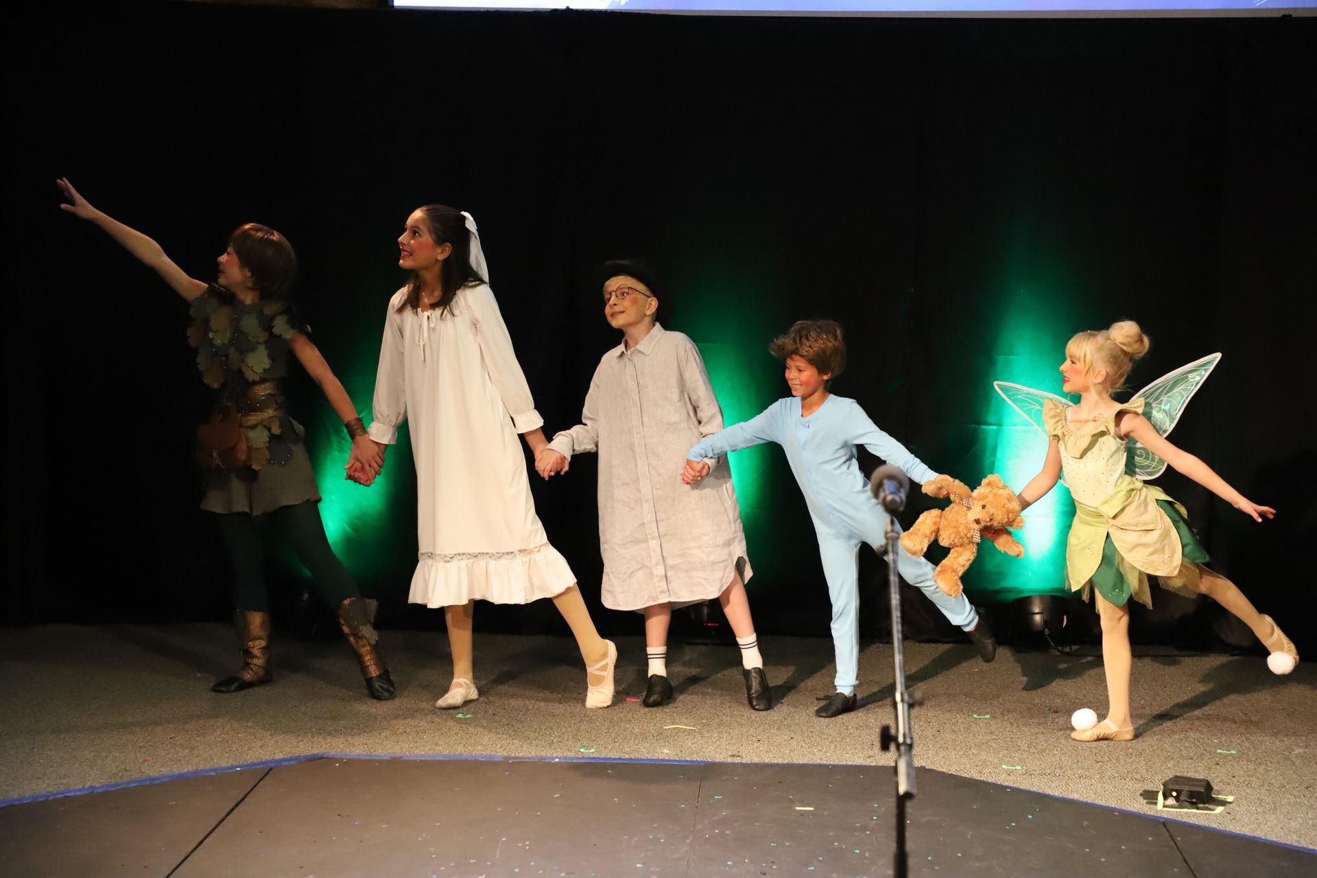 A group of children dressed as peter pan and tinkerbell are holding hands on a stage.
