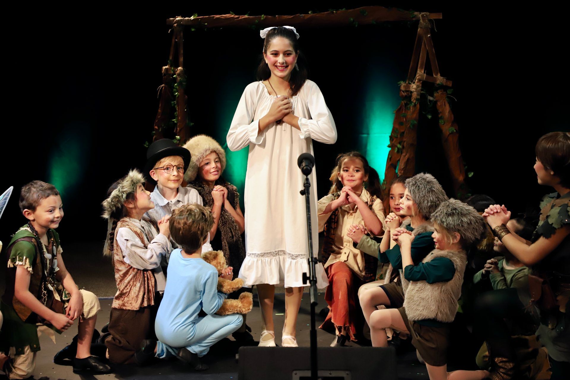 A girl is standing in front of a microphone surrounded by children on a stage.