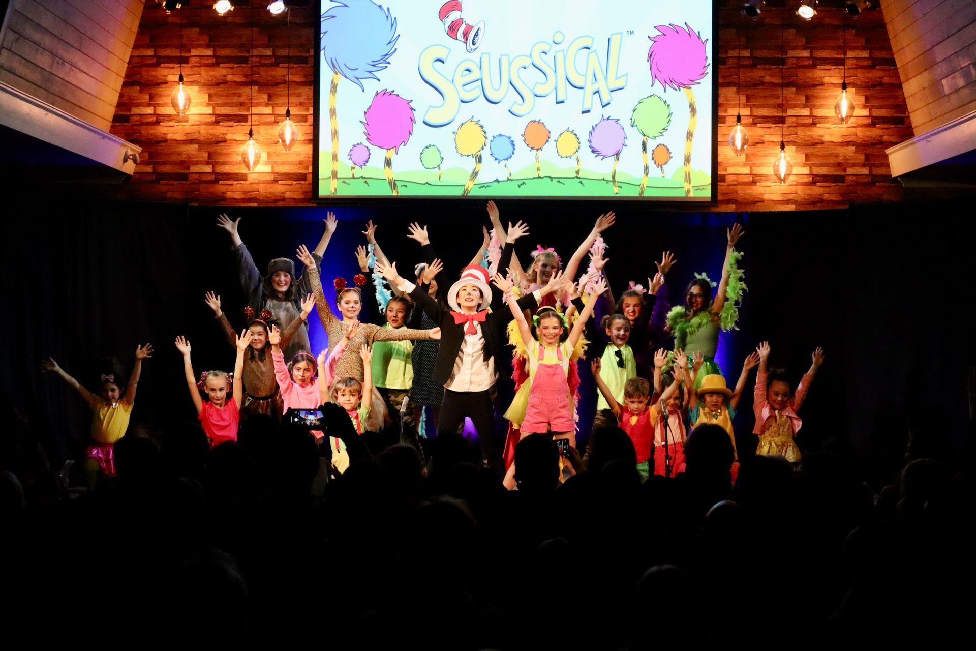 A group of children are dancing on a stage in front of a large screen.