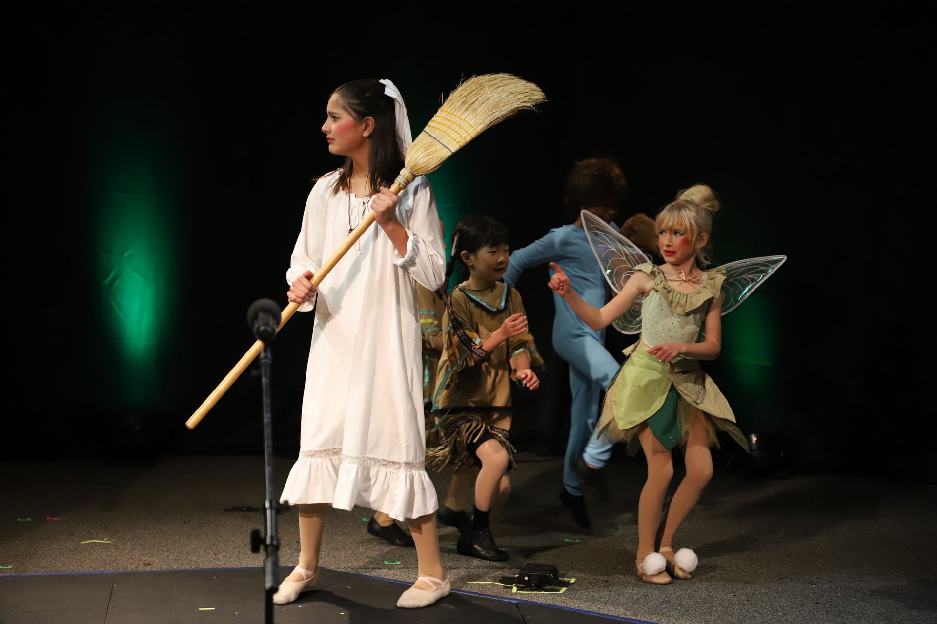 A girl in a white dress is holding a broom in front of a microphone.
