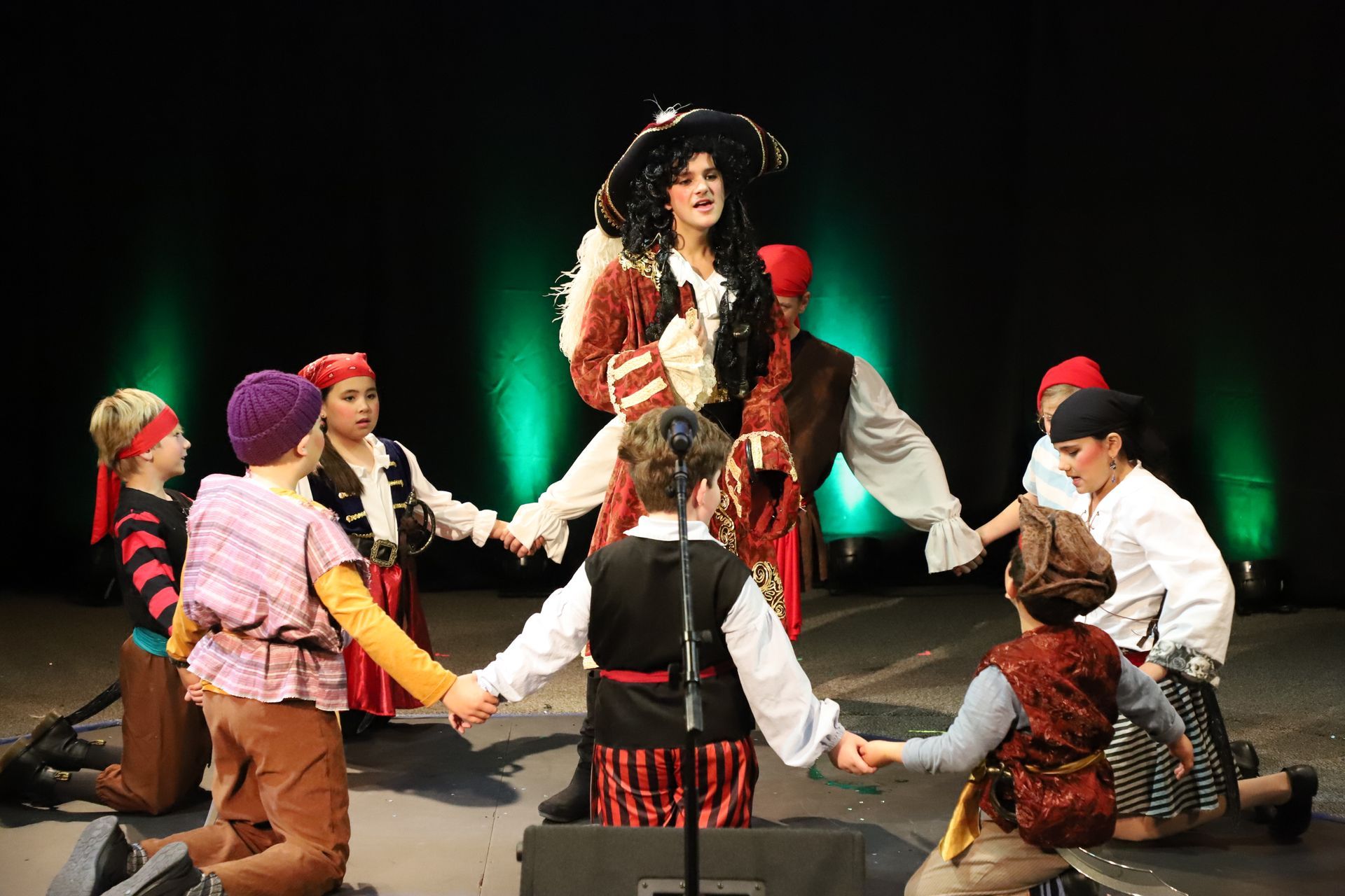 A group of children dressed in pirate costumes are holding hands on a stage.