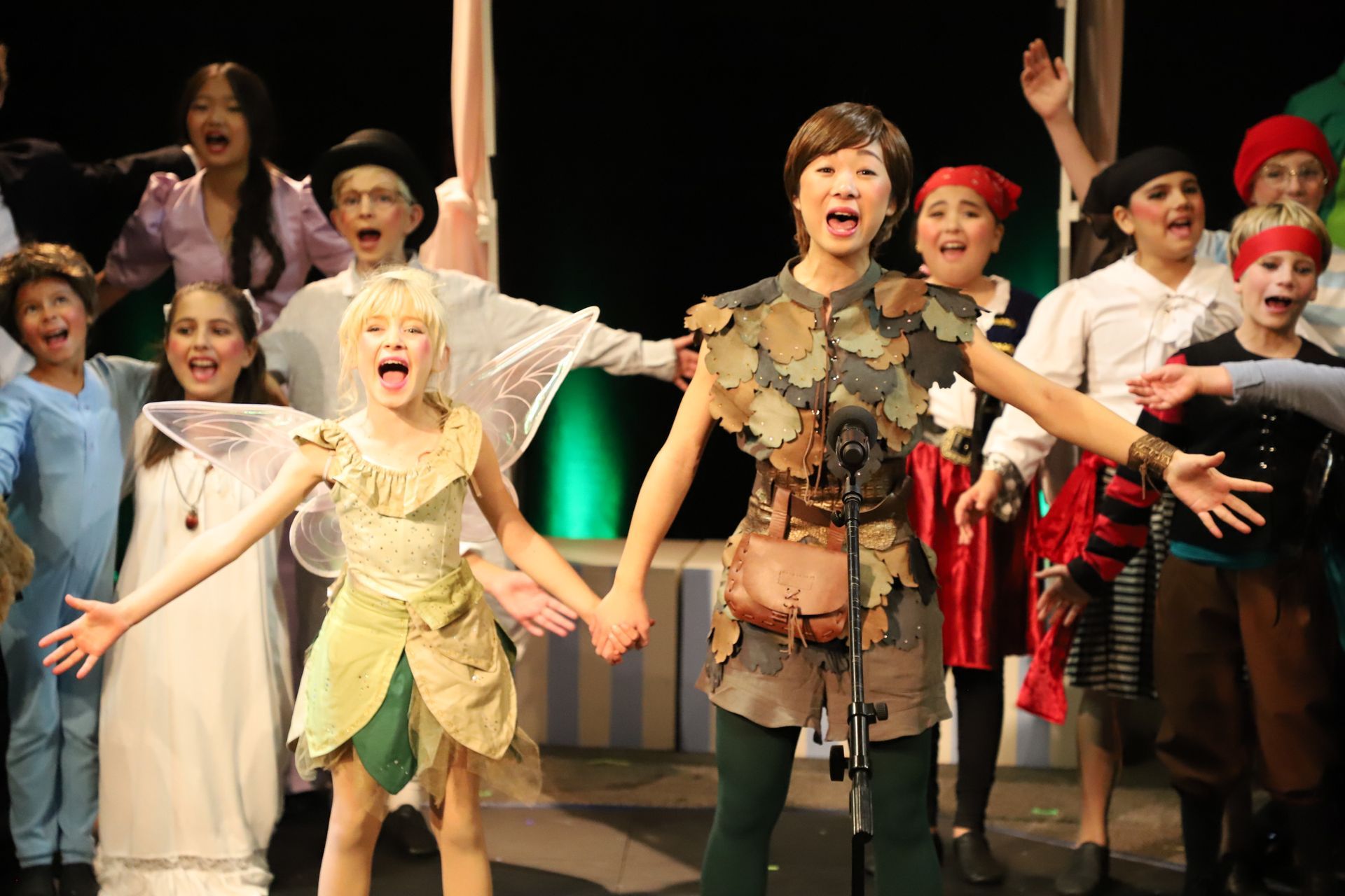 A group of children dressed as peter pan and tinkerbell are holding hands on a stage.