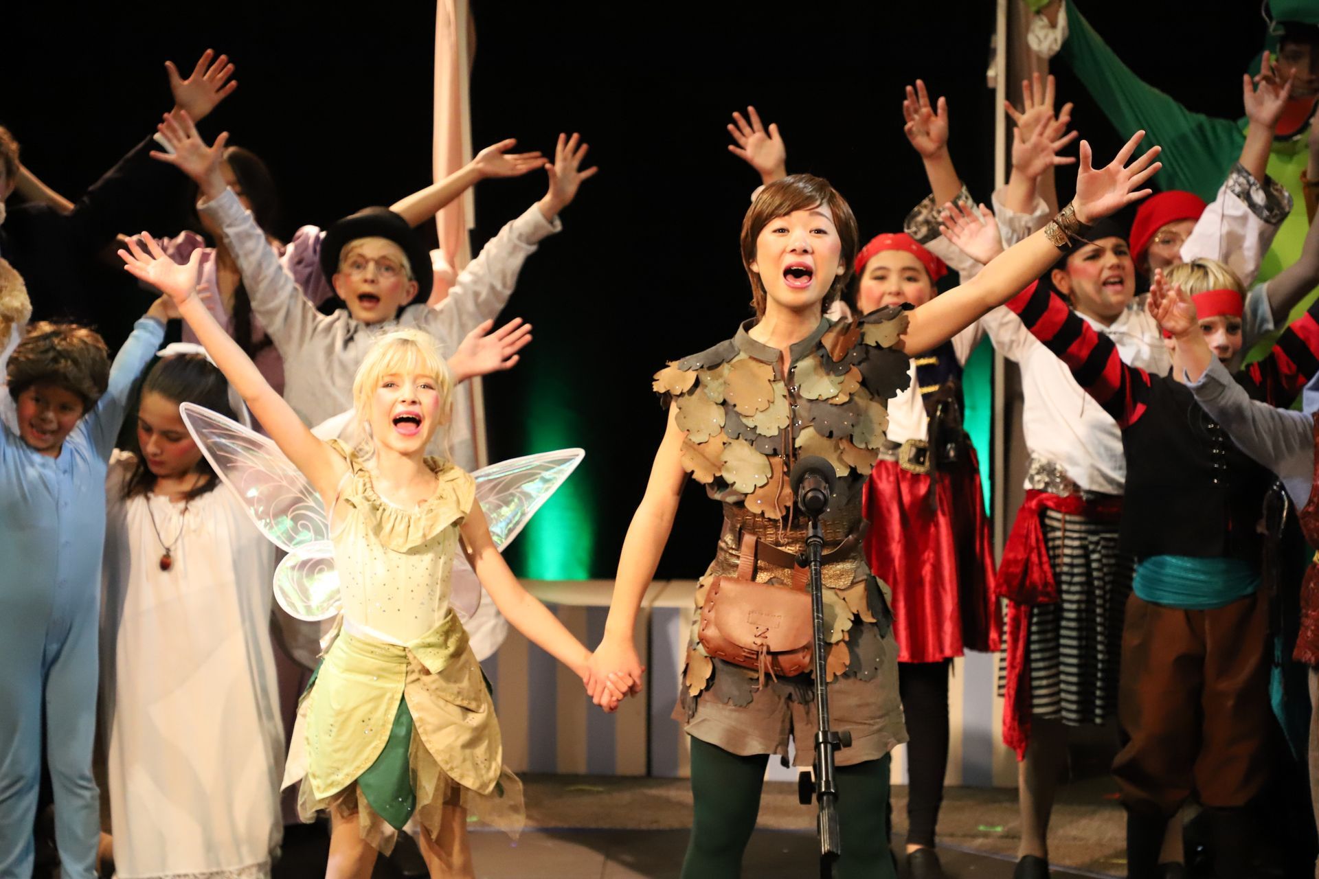 A group of children dressed as peter pan and tinkerbell are holding hands on a stage.