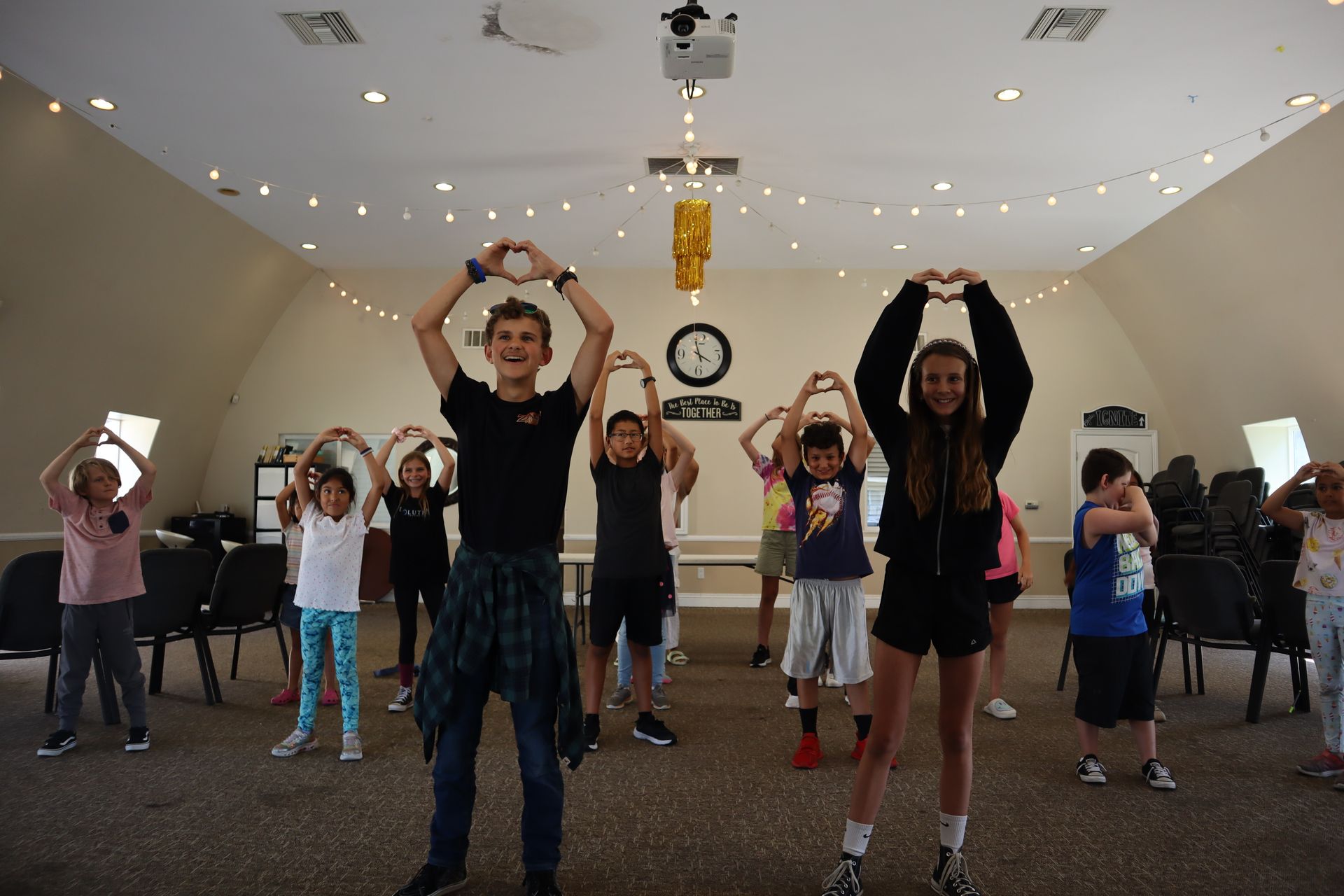 A group of children are standing in a room making heart shapes with their hands.