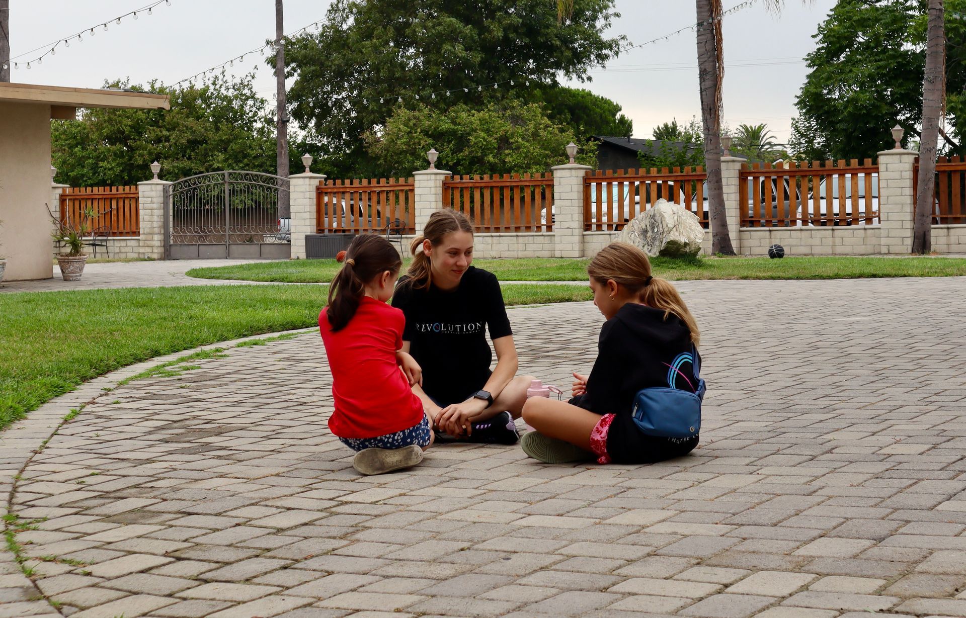 Three young girls are sitting on a brick driveway holding hands