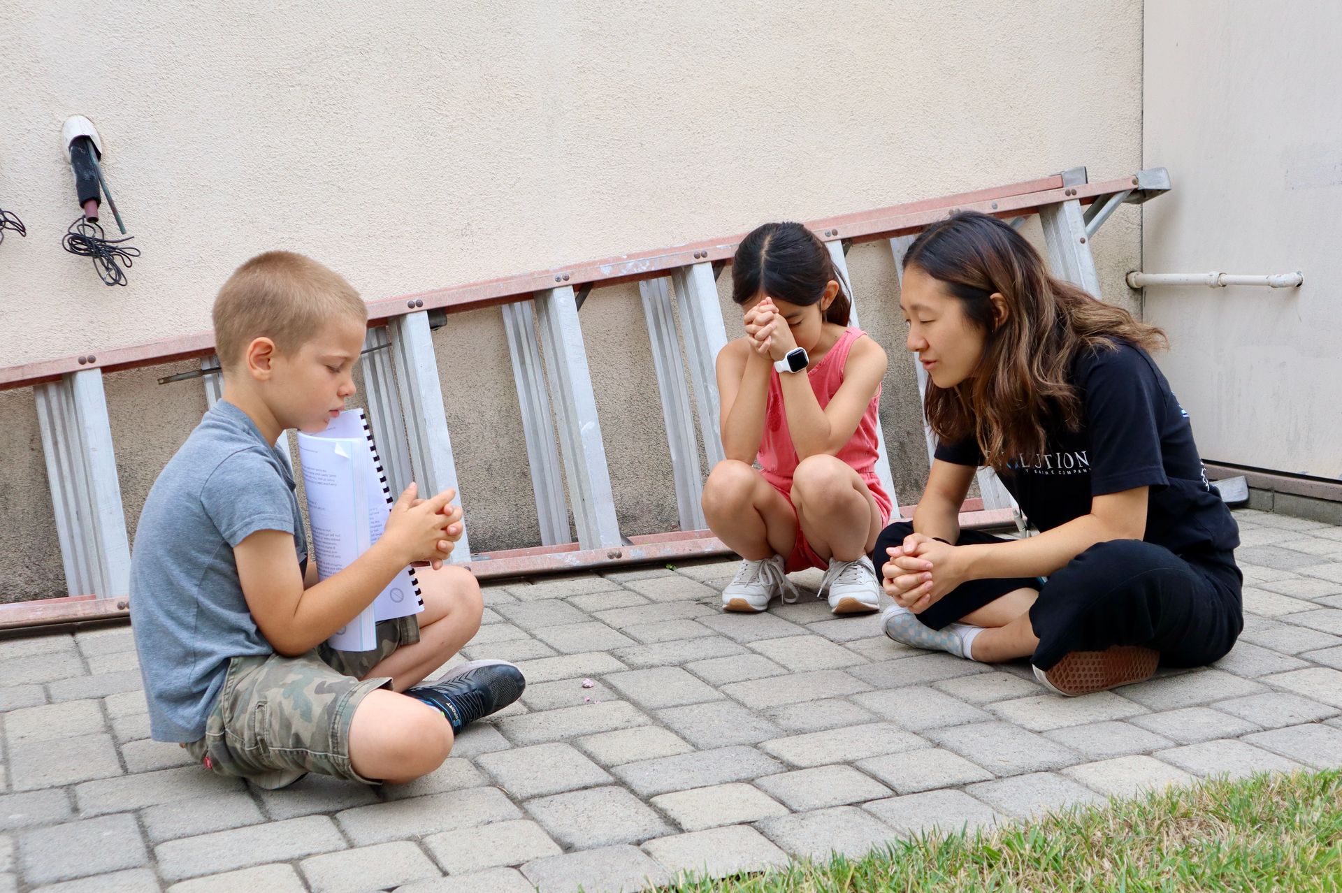 A boy and two girls are sitting on a brick sidewalk in front of a ladder.