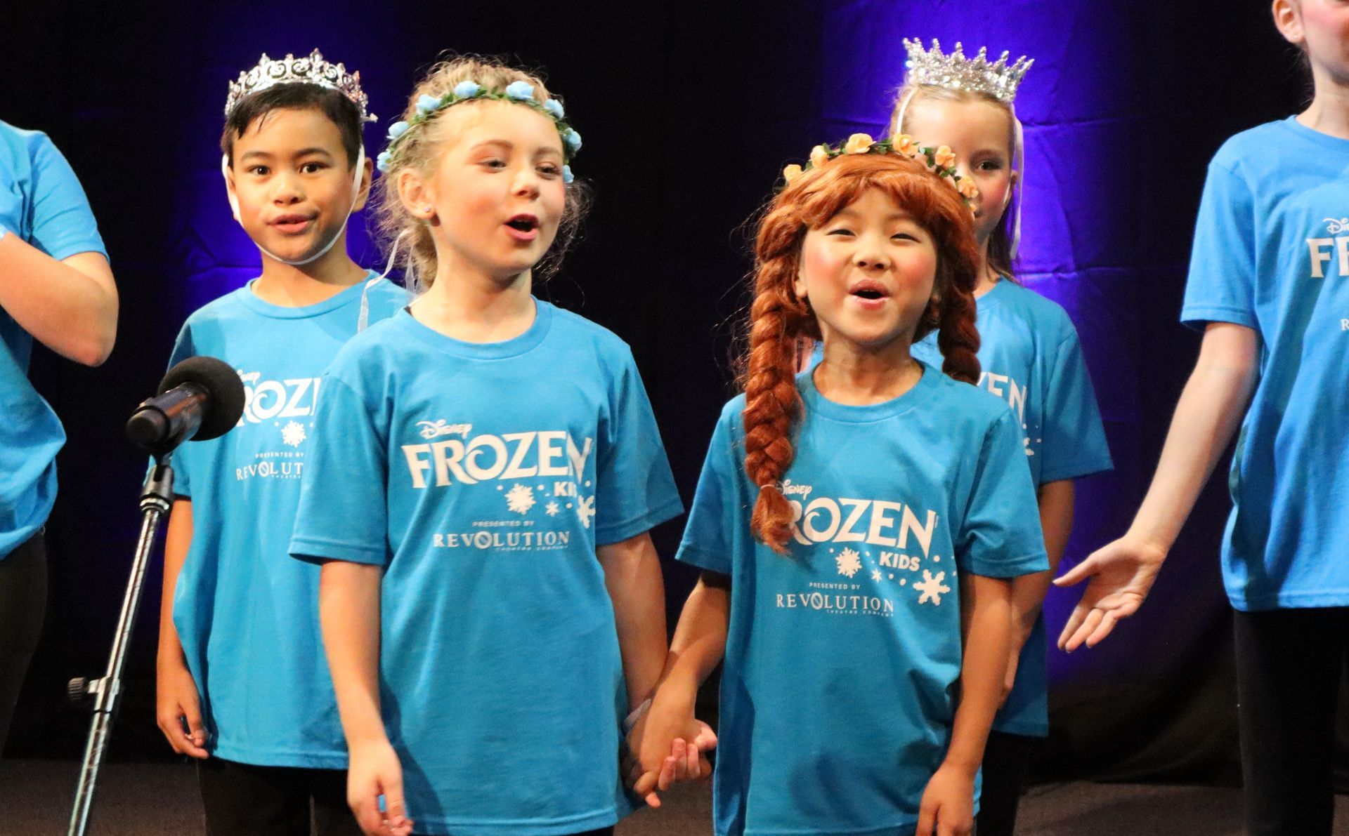 A group of children wearing blue frozen shirts are singing into microphones.