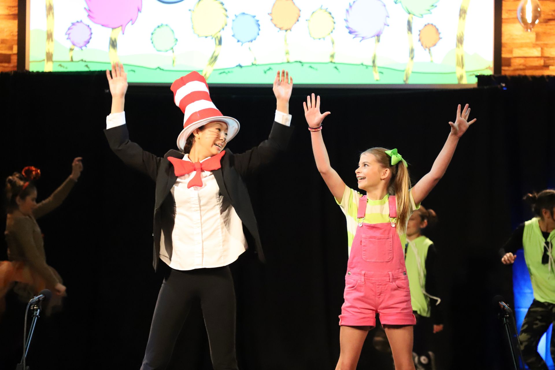 A man in a cat in the hat and a girl in pink overalls are dancing on a stage.