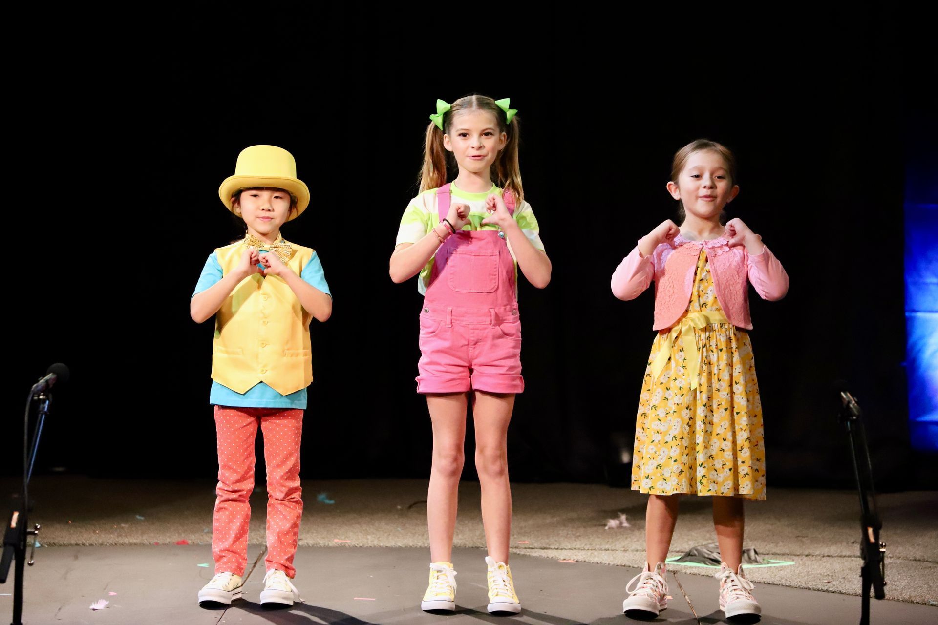Three children are standing on a stage in front of microphones.