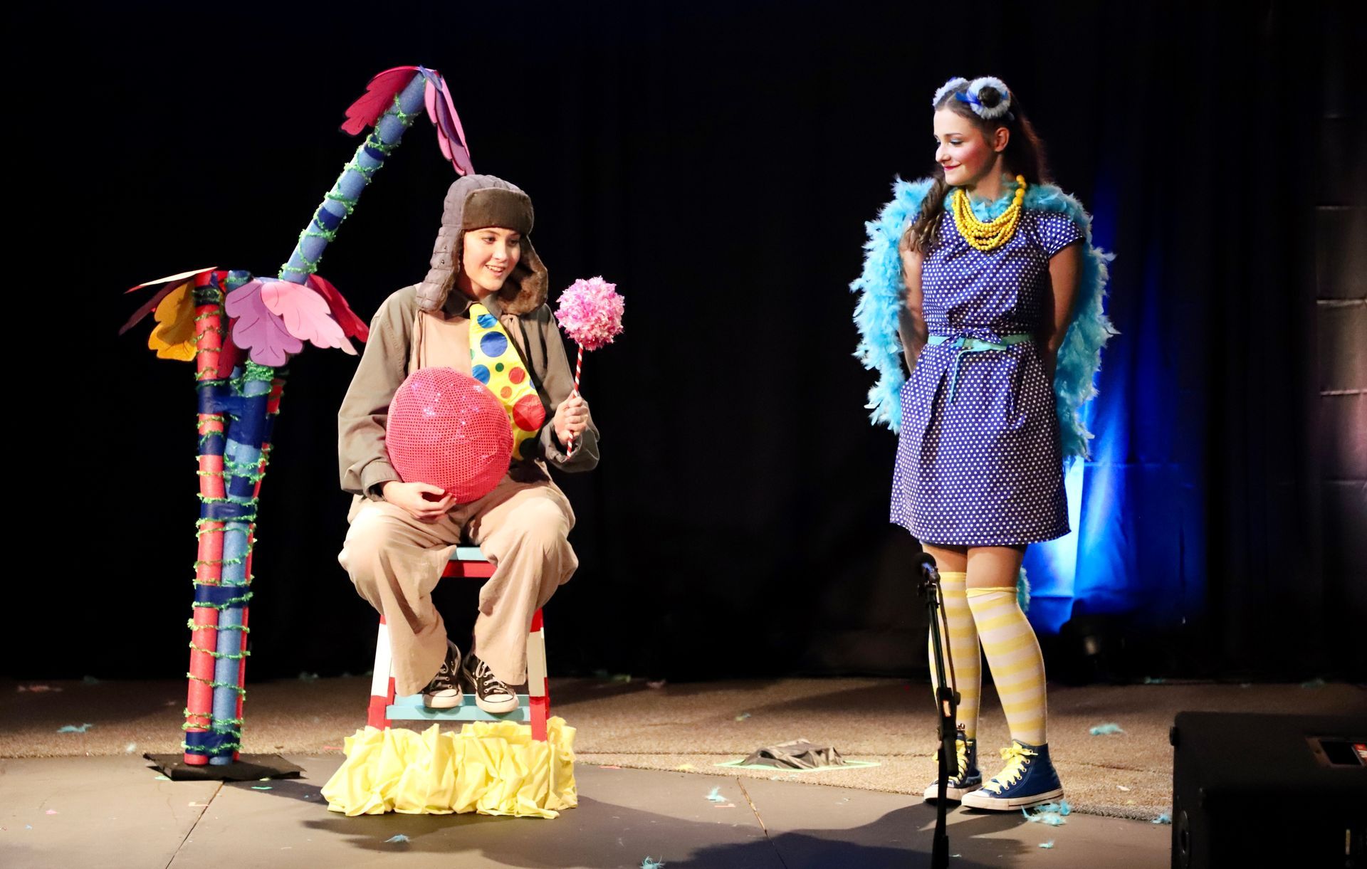 A man is sitting on a chair while a woman stands next to him on a stage.