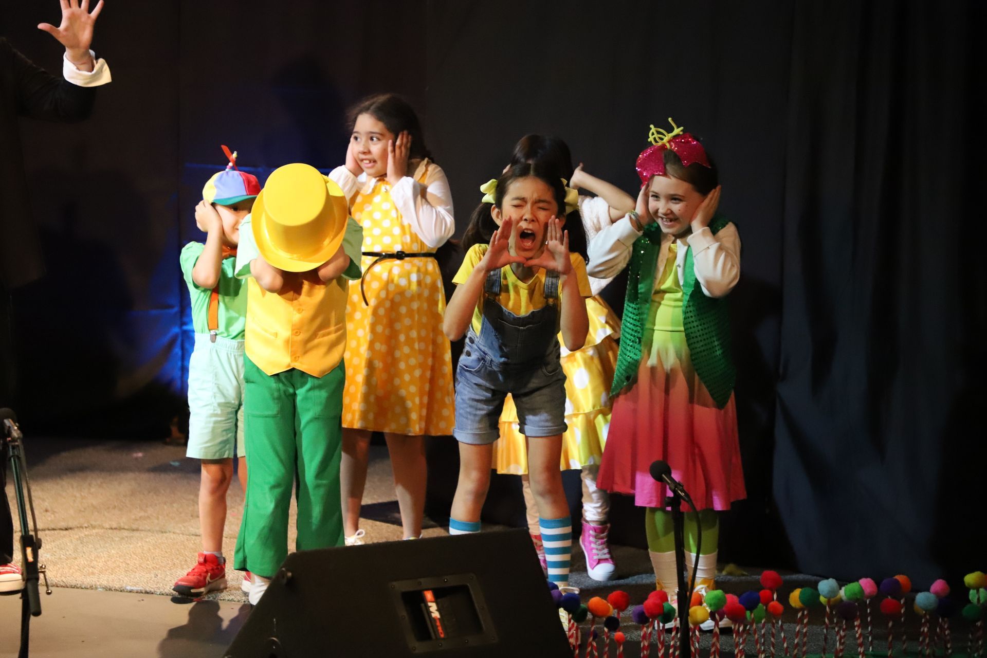 A group of children are standing on a stage covering their ears with their hands.