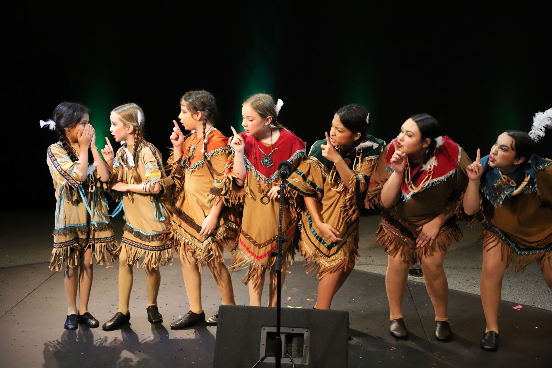 A group of young girls in native american costumes are singing into microphones on a stage.