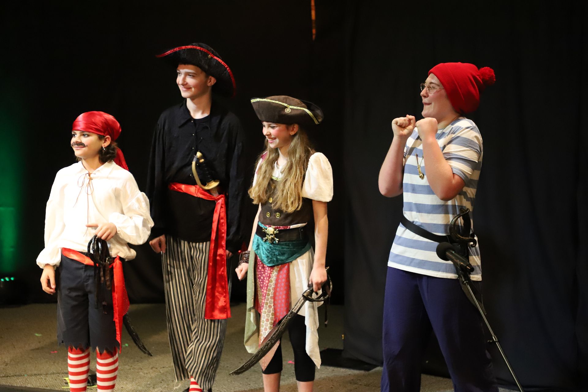 A group of children dressed in pirate costumes are standing on a stage holding swords.