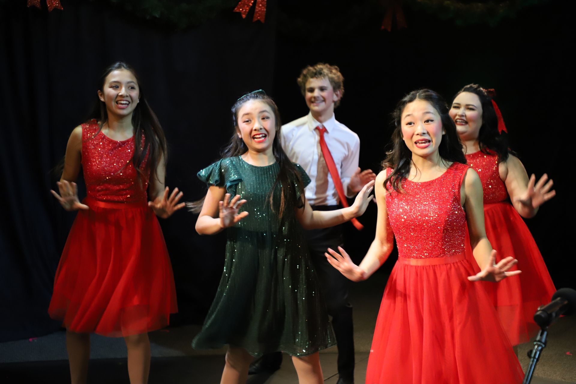 A group of young girls in red dresses are singing on a stage.